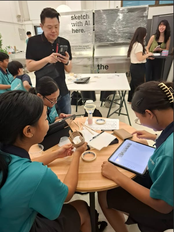 Students in teal uniforms work at a round table with craft supplies and a tablet at an AI sketching workshop.