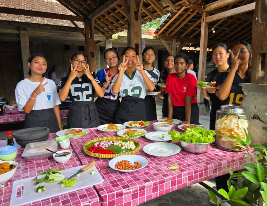 Eight young people in aprons pose with peace signs behind a table filled with colorful food dishes outdoors.