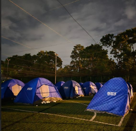 Several blue Adventure World dome tents set up on a grass field at dusk with dark cloudy sky and trees behind.