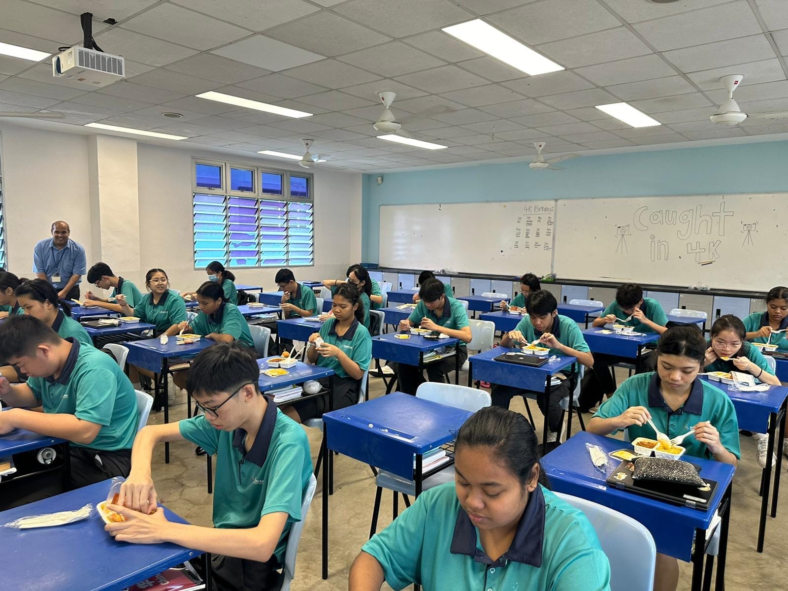 Students in teal uniforms eating meals at blue desks in a classroom with a whiteboard reading "Caught in 4K."