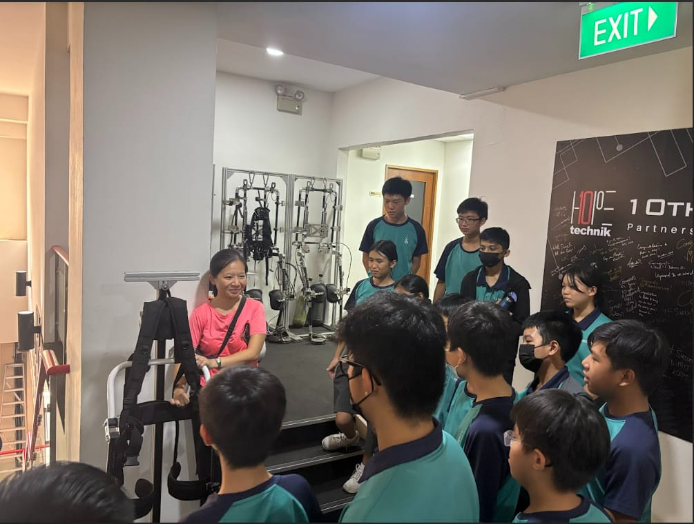 A woman in a pink shirt presents rehabilitation equipment to a group of students in teal uniforms indoors.