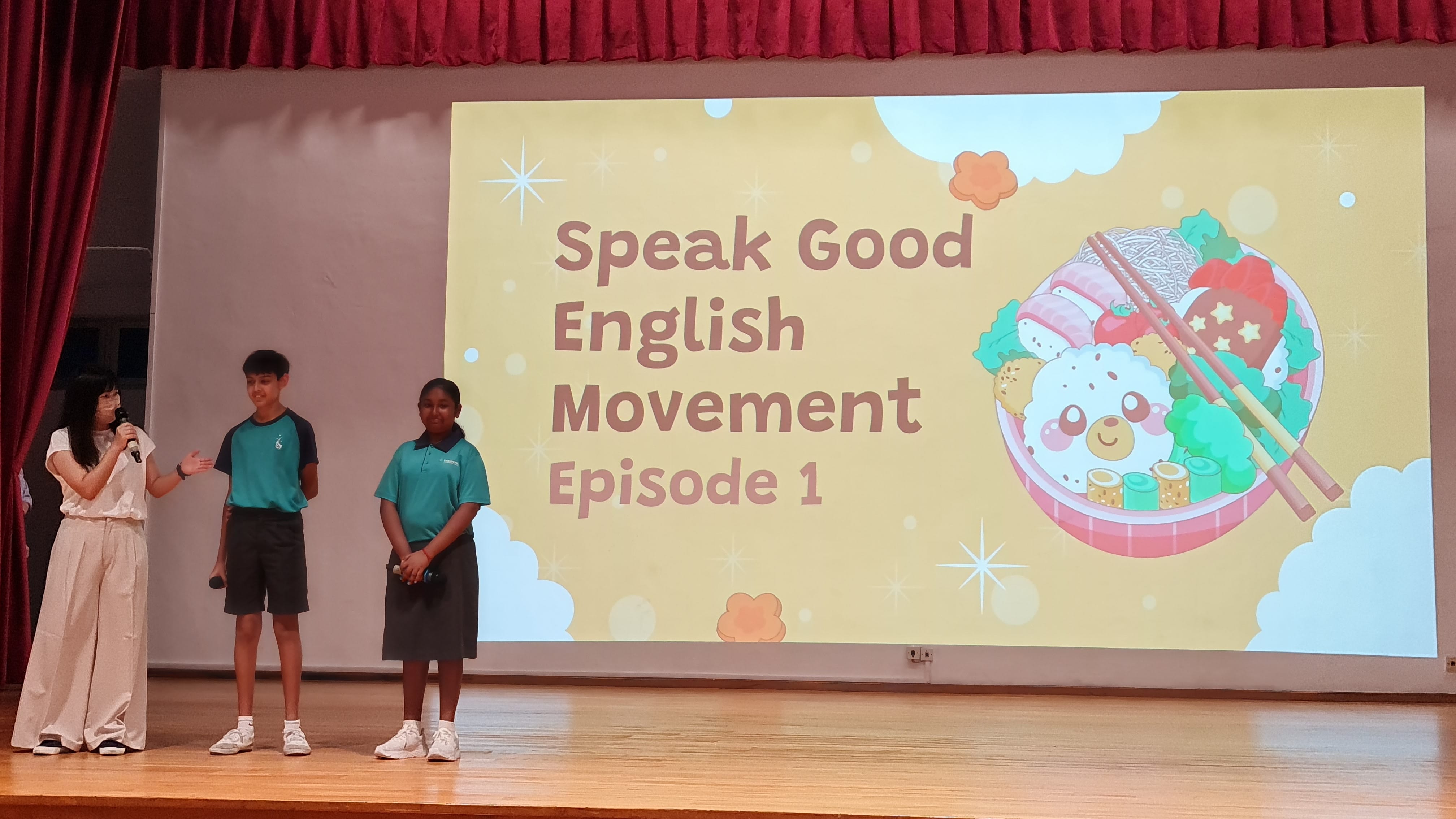 Three people stand on a stage beside a projected slide reading Speak Good English Movement Episode 1.