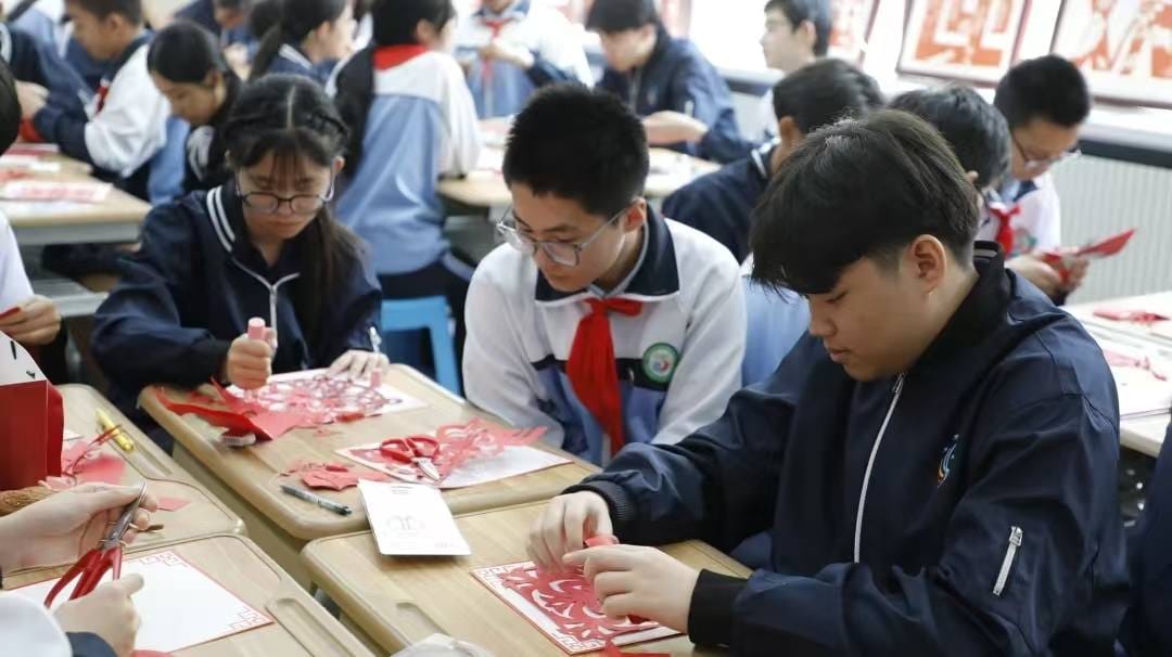 Students in school uniforms practice traditional Chinese paper cutting with red paper and scissors at classroom desks.