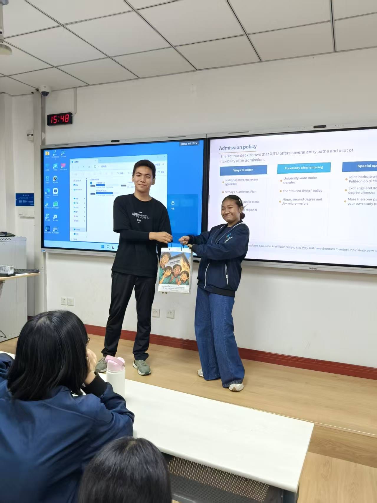 Two students stand at the front of a classroom exchanging a gift bag in front of dual presentation screens.