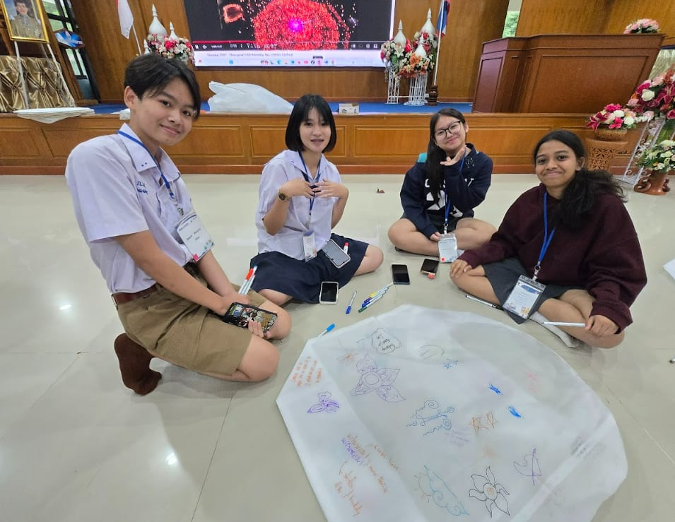 Four students wearing lanyards sit on a floor around a large paper with colorful drawings and writing.