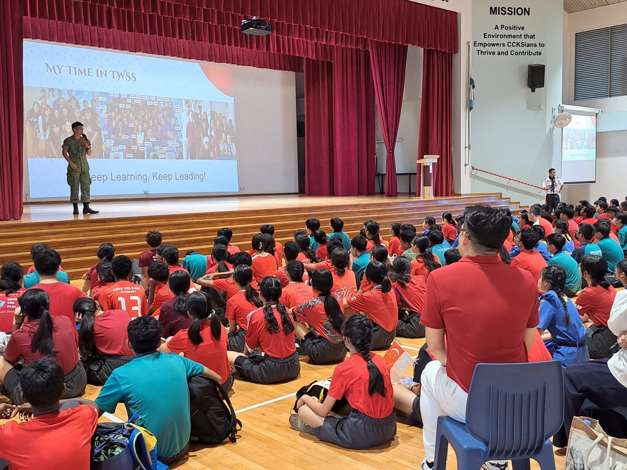 Students in red shirts sit on a hall floor watching a speaker on stage with a projected slide reading "Keep Learning, Keep Leading."