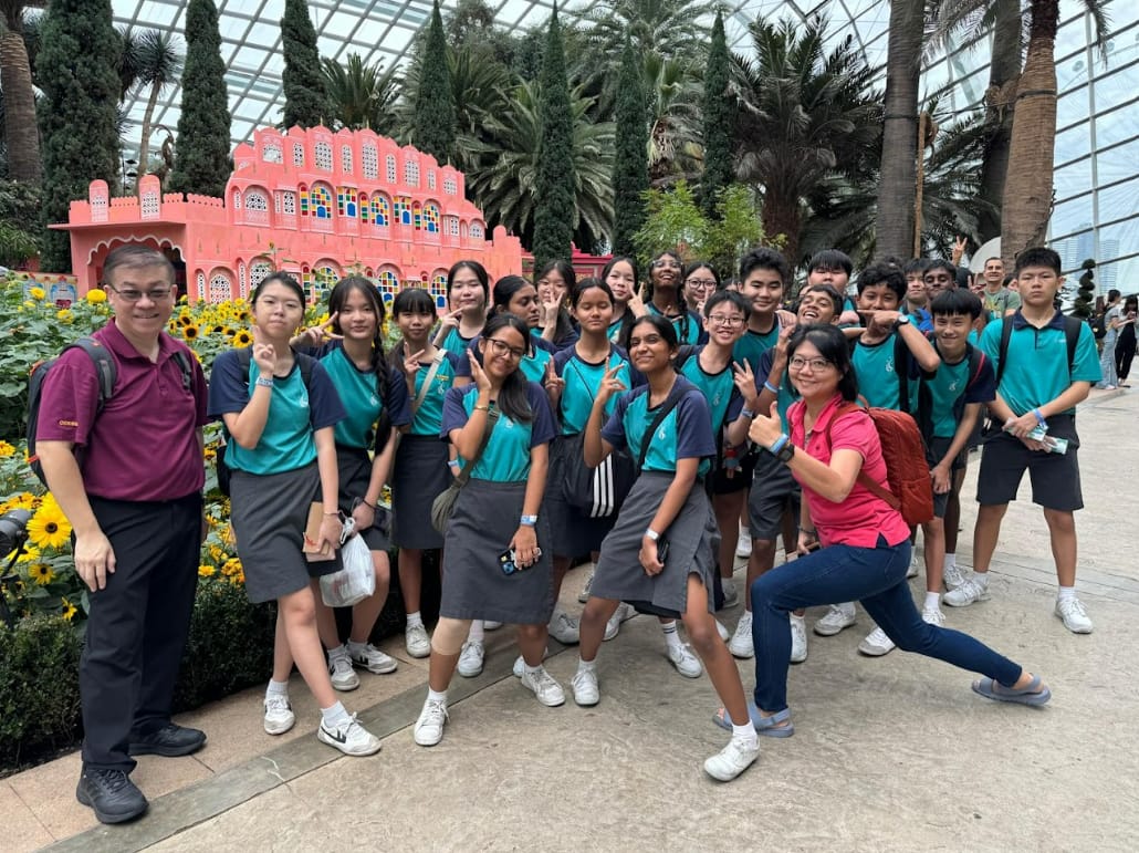 A group of students in teal uniforms pose with two adults inside a greenhouse near a pink palace replica and sunflowers.