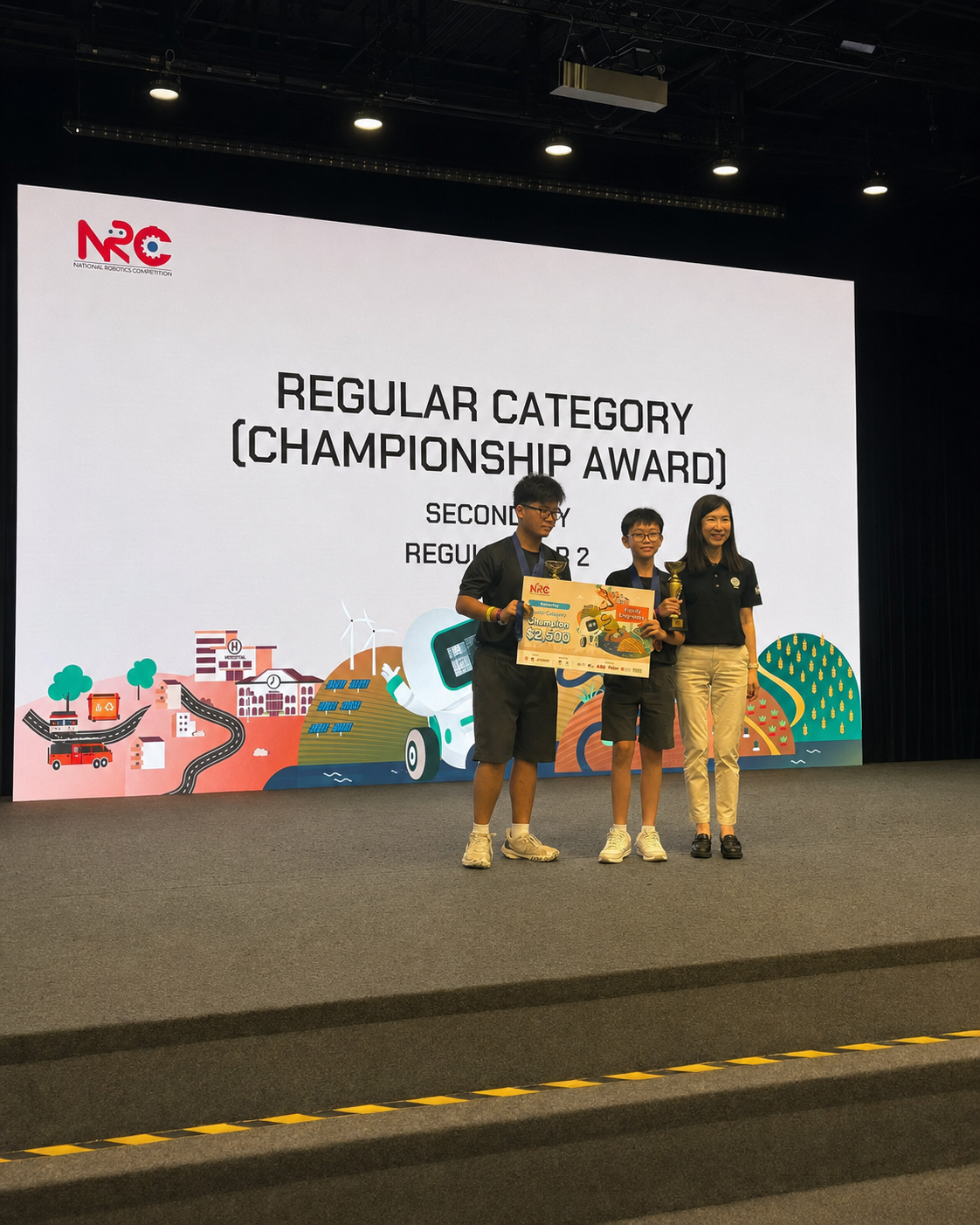 Two boys hold a champion cheque and trophy on stage at an NRC Regular Category Championship Award ceremony.