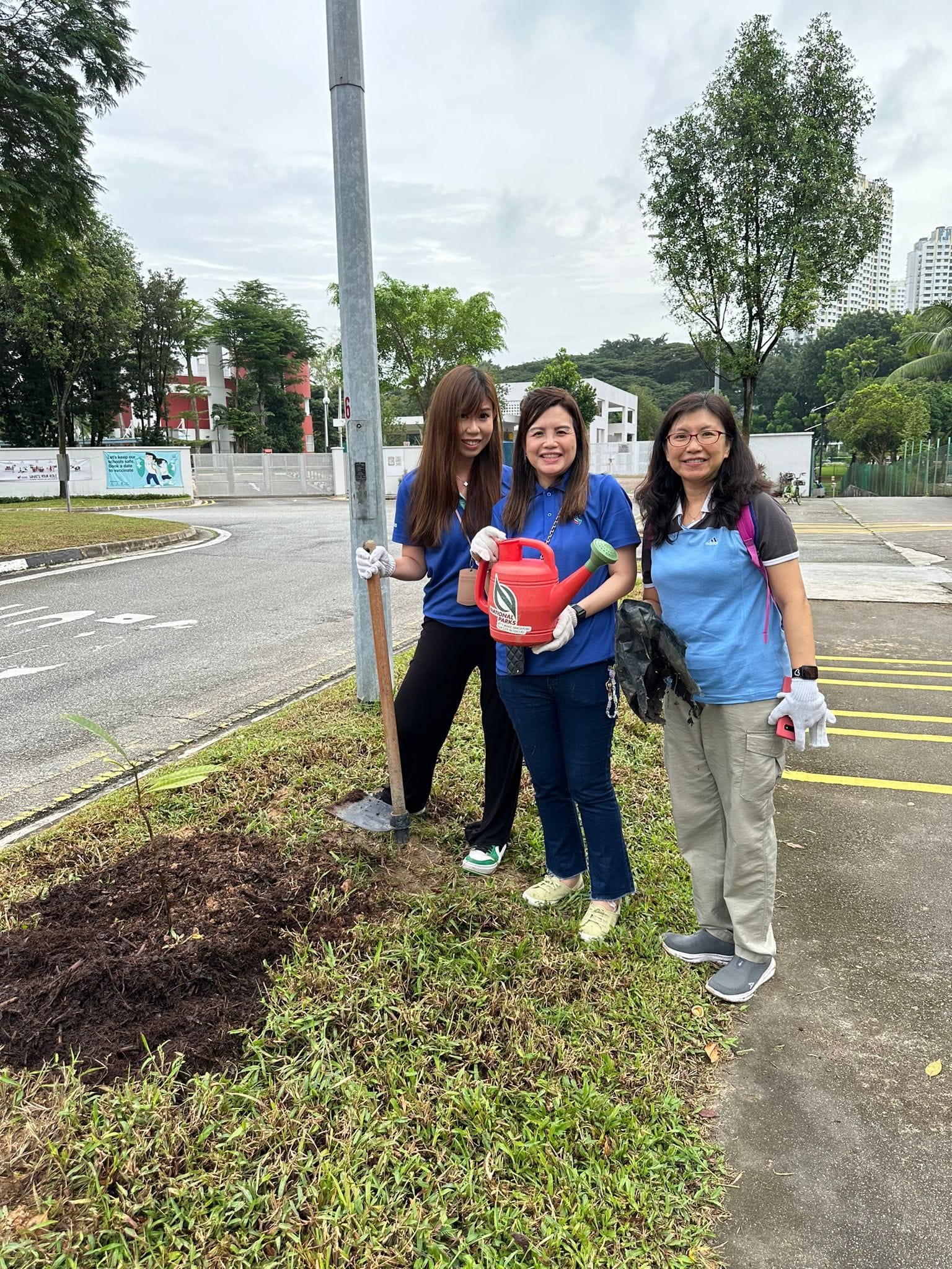 Three women stand on a grass patch near a road, one holding a red watering can and another holding a shovel.