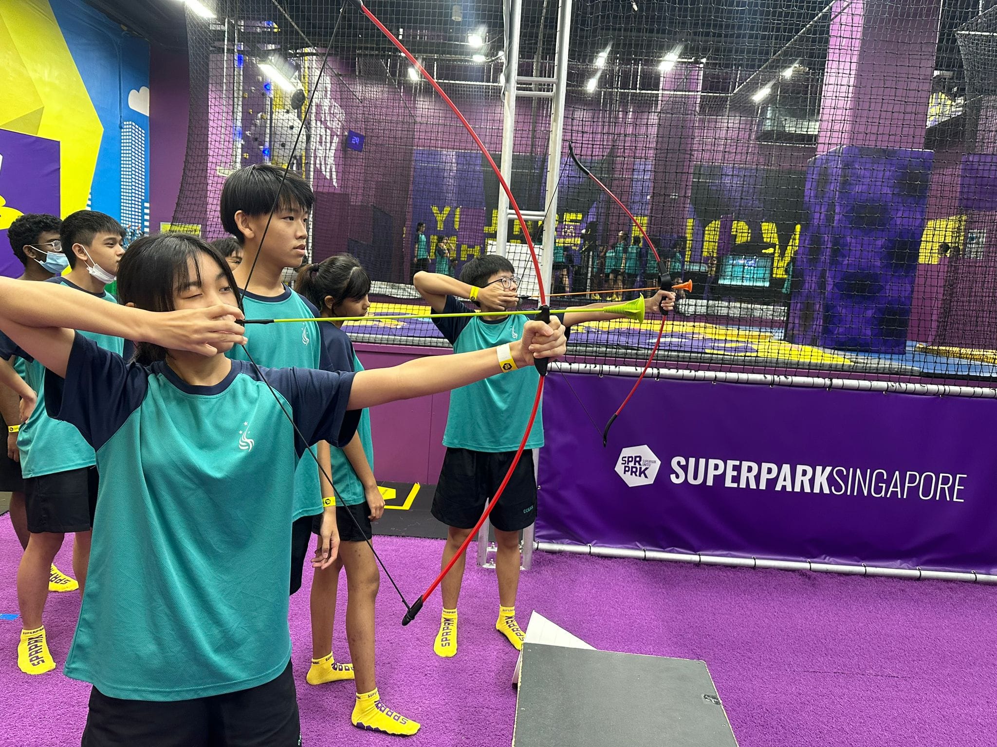 Students in teal uniforms practice archery with red bows at SuperPark Singapore indoor activity center.