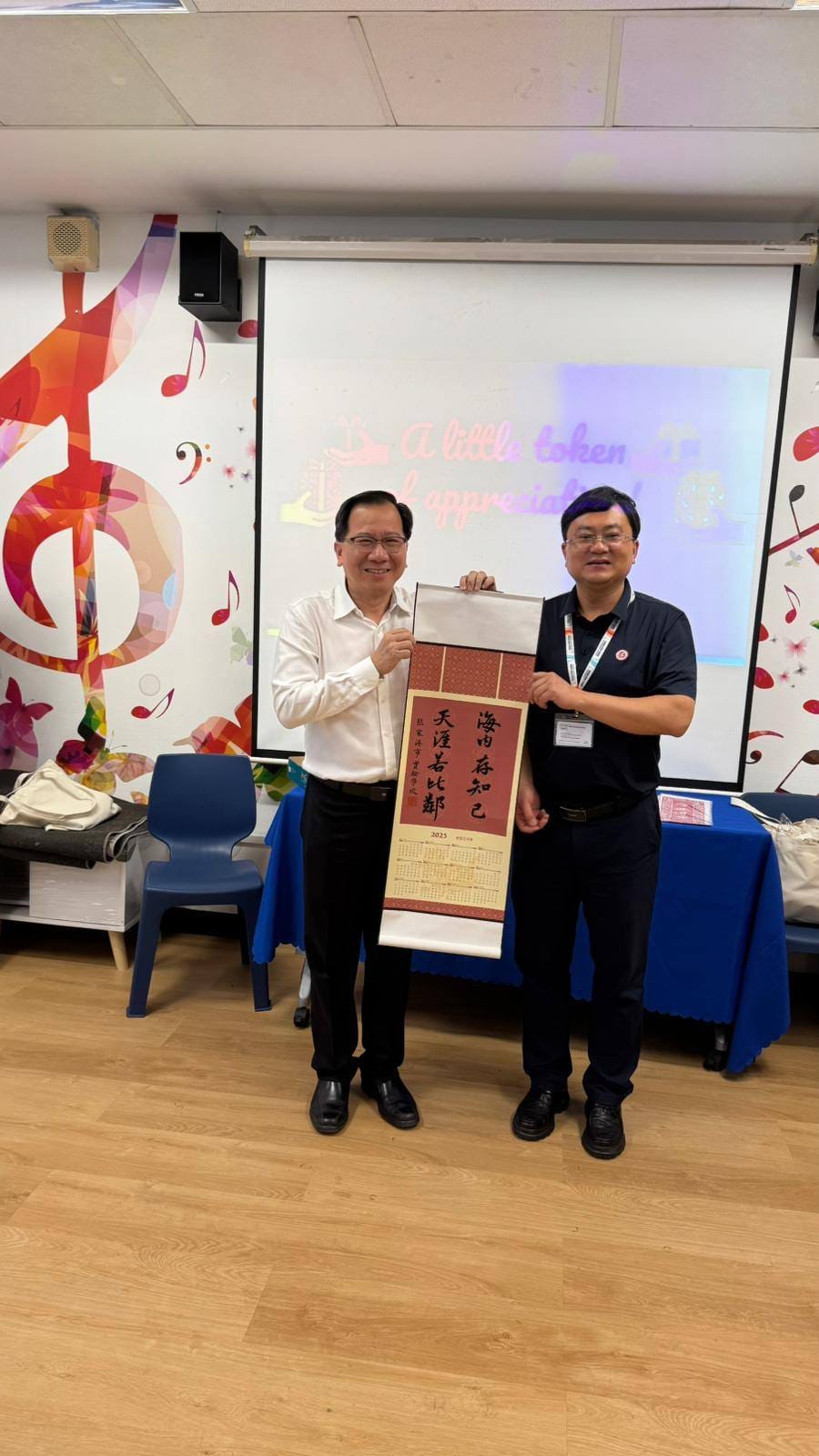 Two men stand together holding a Chinese calligraphy scroll with a 2025 calendar in a decorated room.