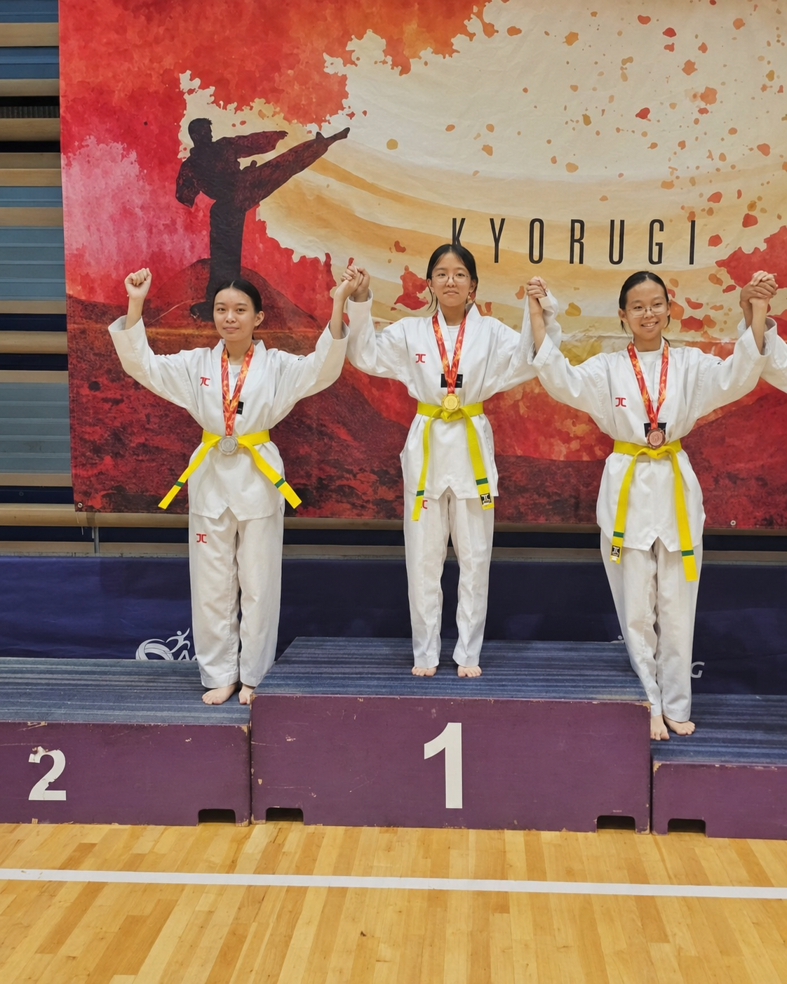 three girls in taekwondo outfits, hold up hands on a podium