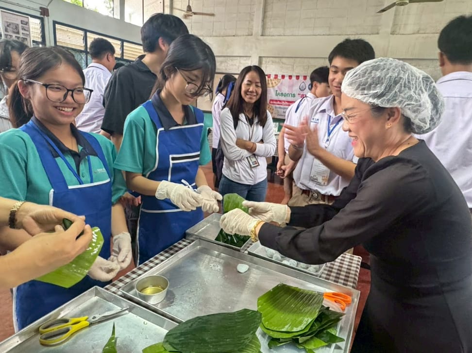 A group of students and an instructor wearing gloves work together wrapping food in banana leaves at a table.