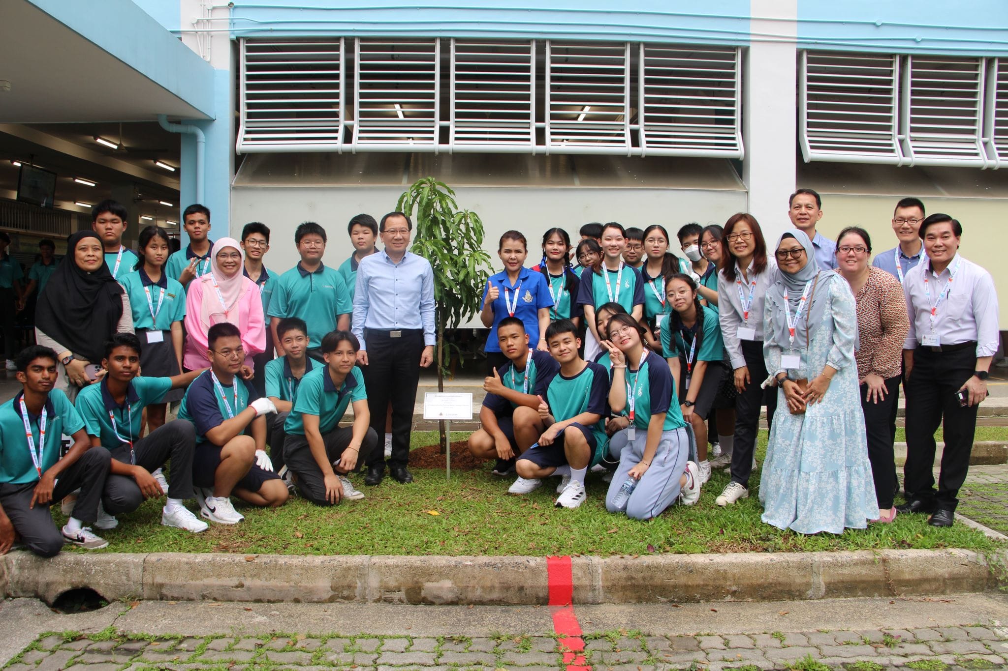 A large group of students and adults in teal uniforms pose around a small newly planted tree outside a school building.