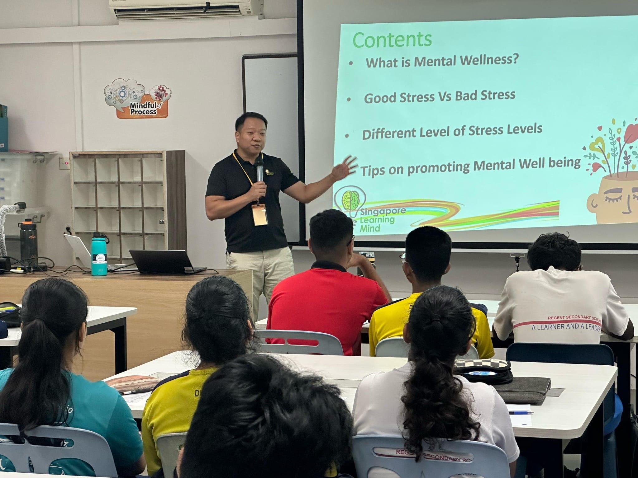 A man with a microphone presents a mental wellness slideshow to students seated at desks in a classroom.