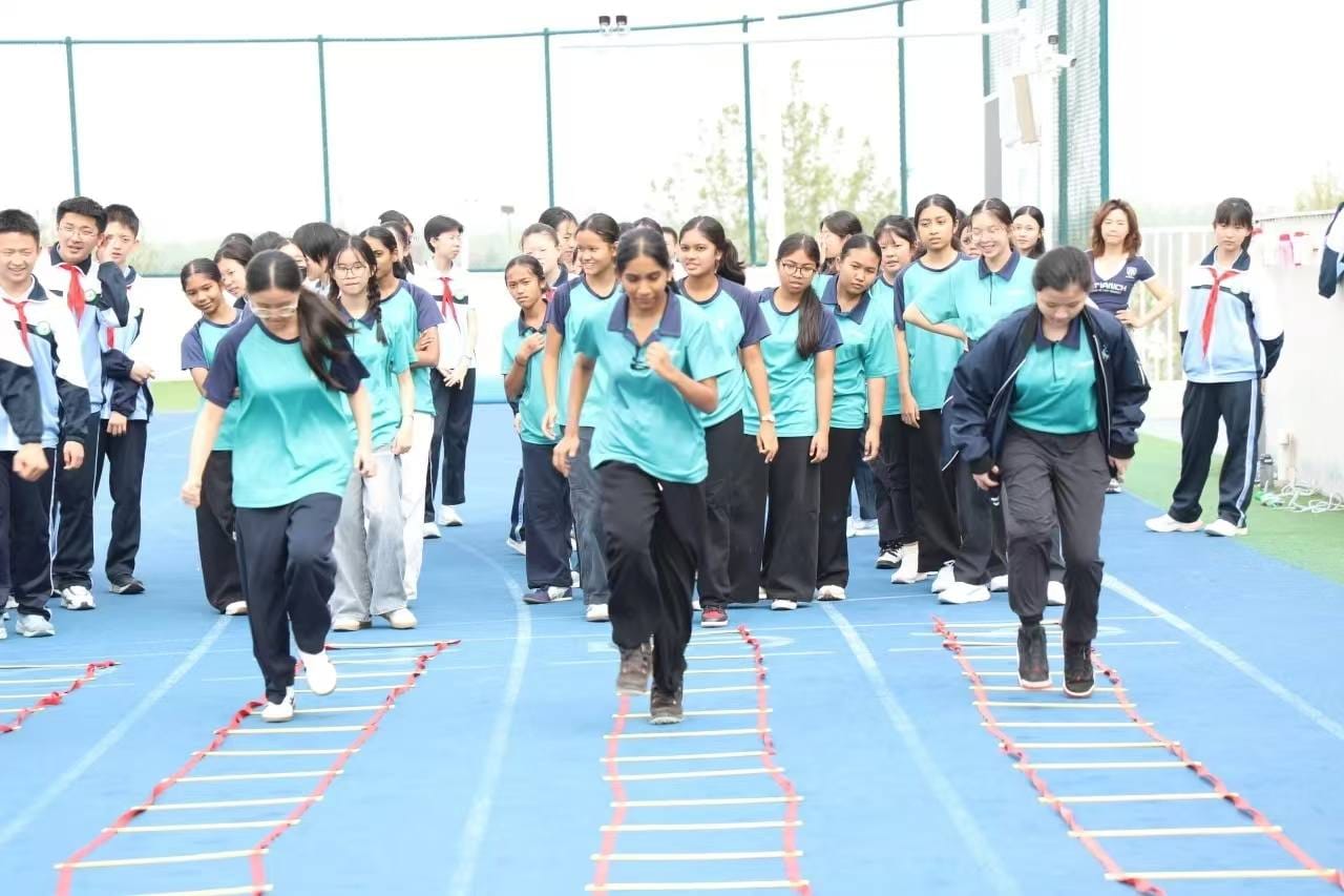 Students in teal shirts stepping through agility ladder drills on a blue outdoor track, with peers watching.