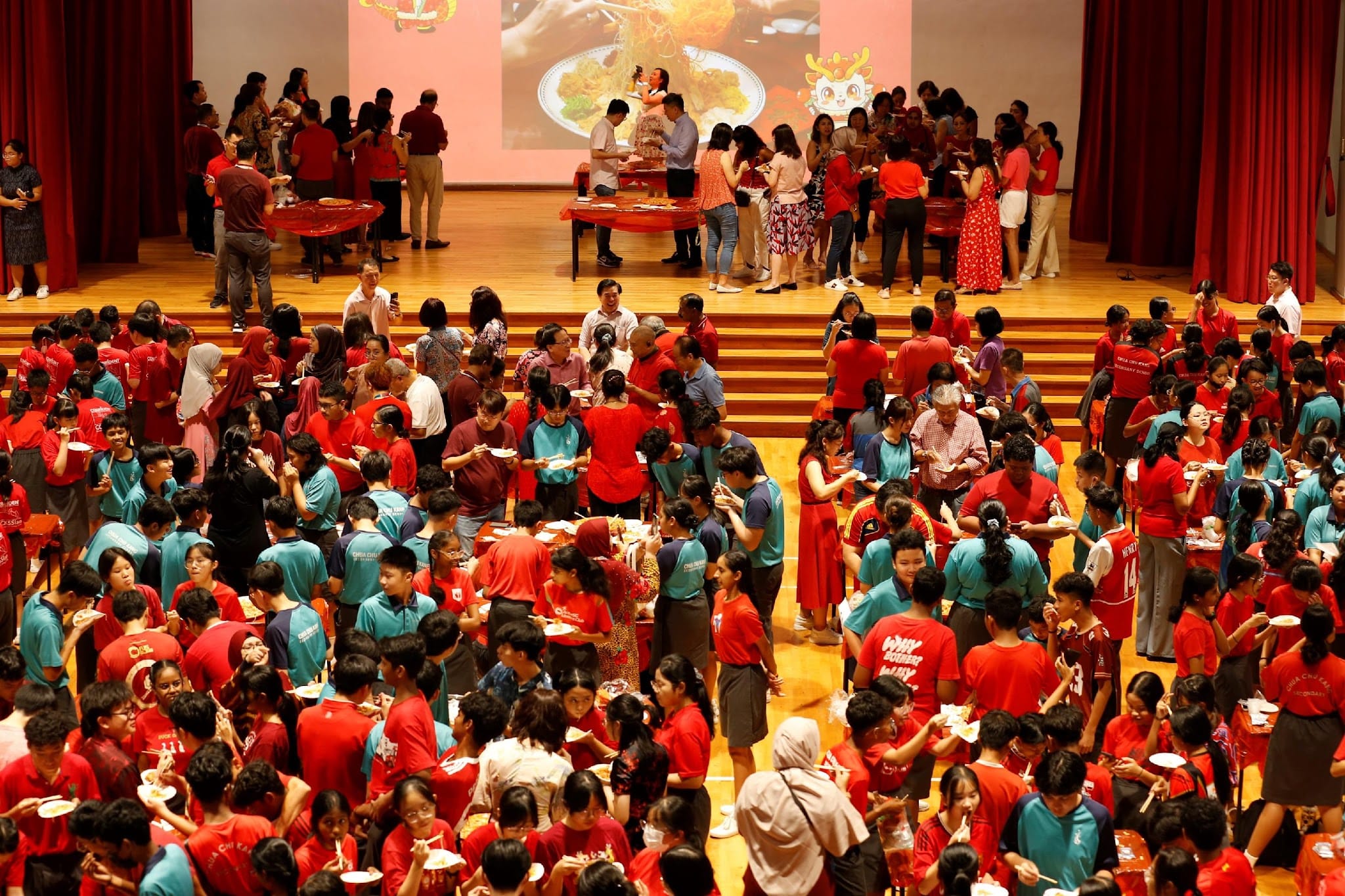 Large crowd in red and teal shirts eating at an indoor celebration with a food projection on stage.