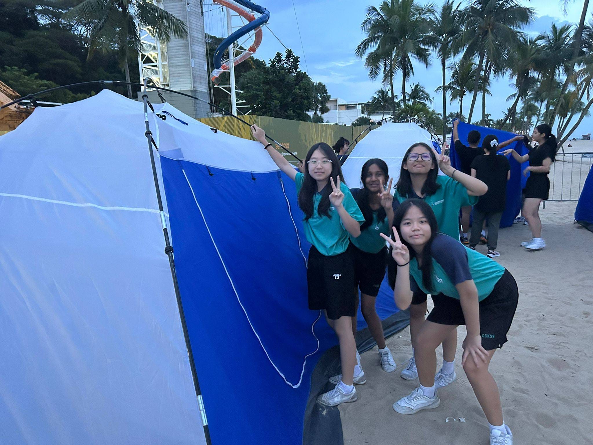Four girls in matching teal uniforms pose together smiling near blue camping tents in an indoor venue.e