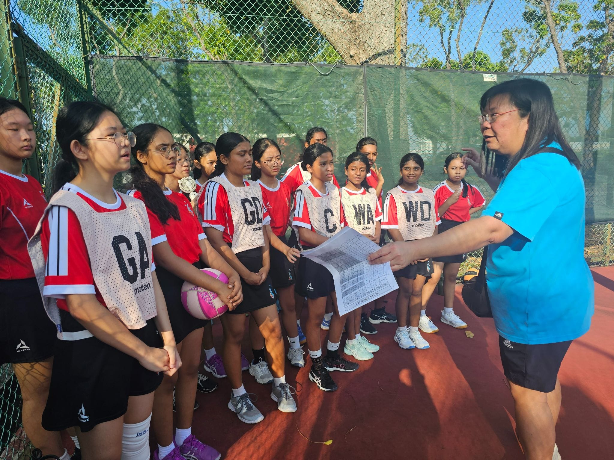 A coach in a blue shirt holds papers while addressing a group of young female netball players wearing position bibs outdoors.