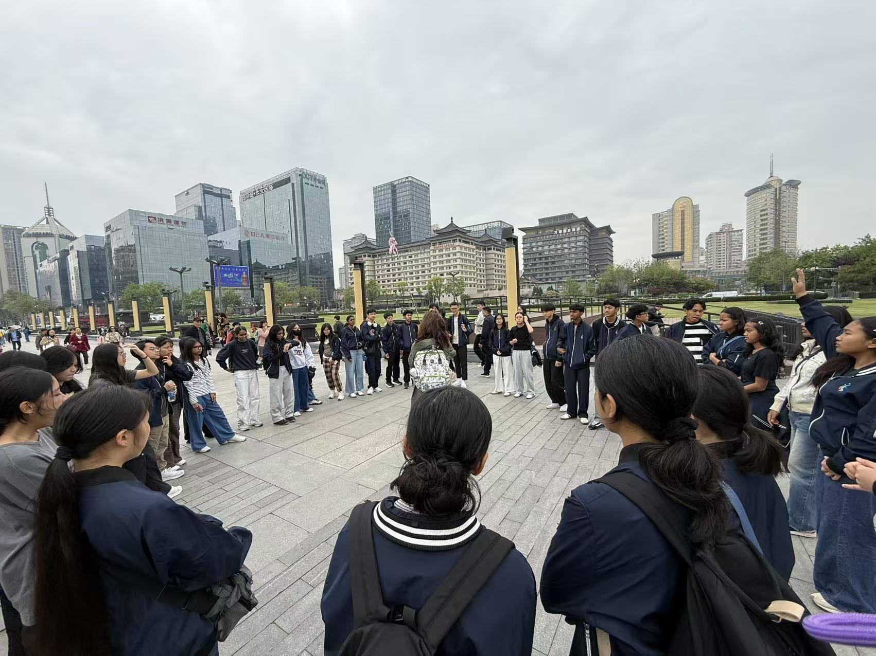 A large group of students in navy uniforms stands in a circle on a city plaza with skyscrapers in the background.