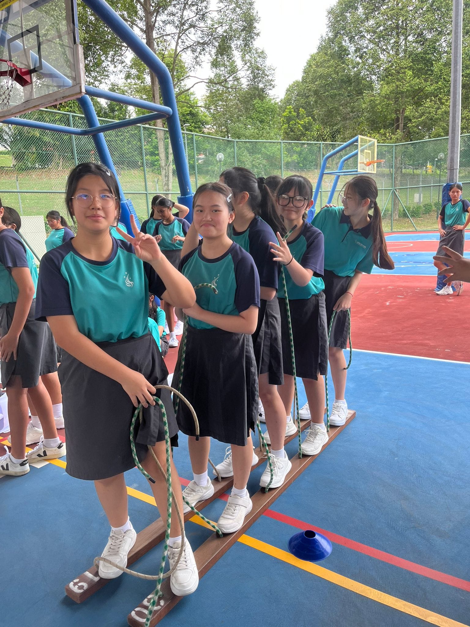 Girls in teal and navy uniforms stand on wooden planks with ropes on a colorful outdoor basketball court.