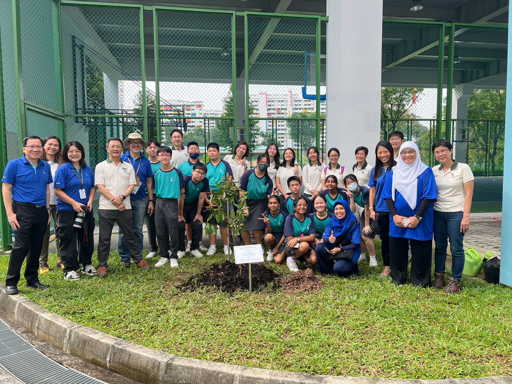 A large group of students and adults pose outdoors around a newly planted small tree on a grass patch.