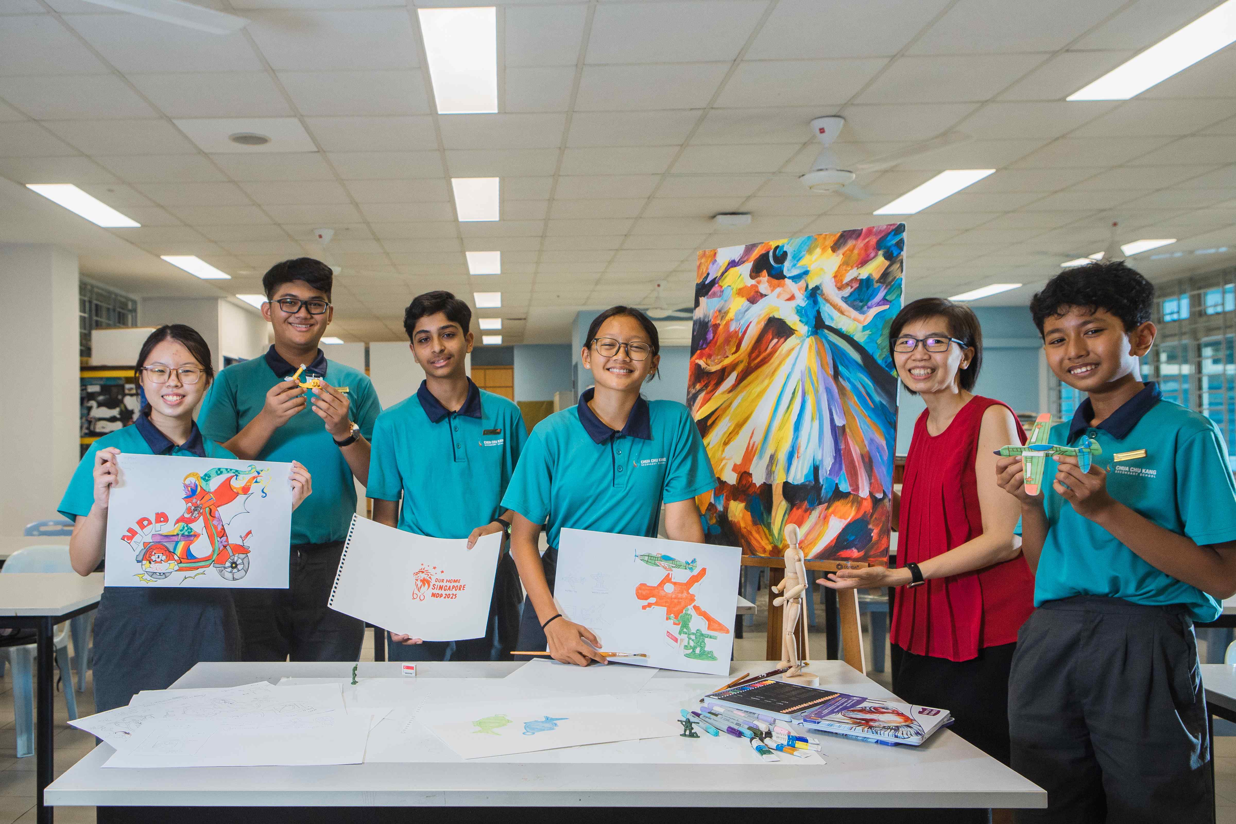 Six students in teal uniforms and a teacher display colorful artwork and small models in a bright classroom.