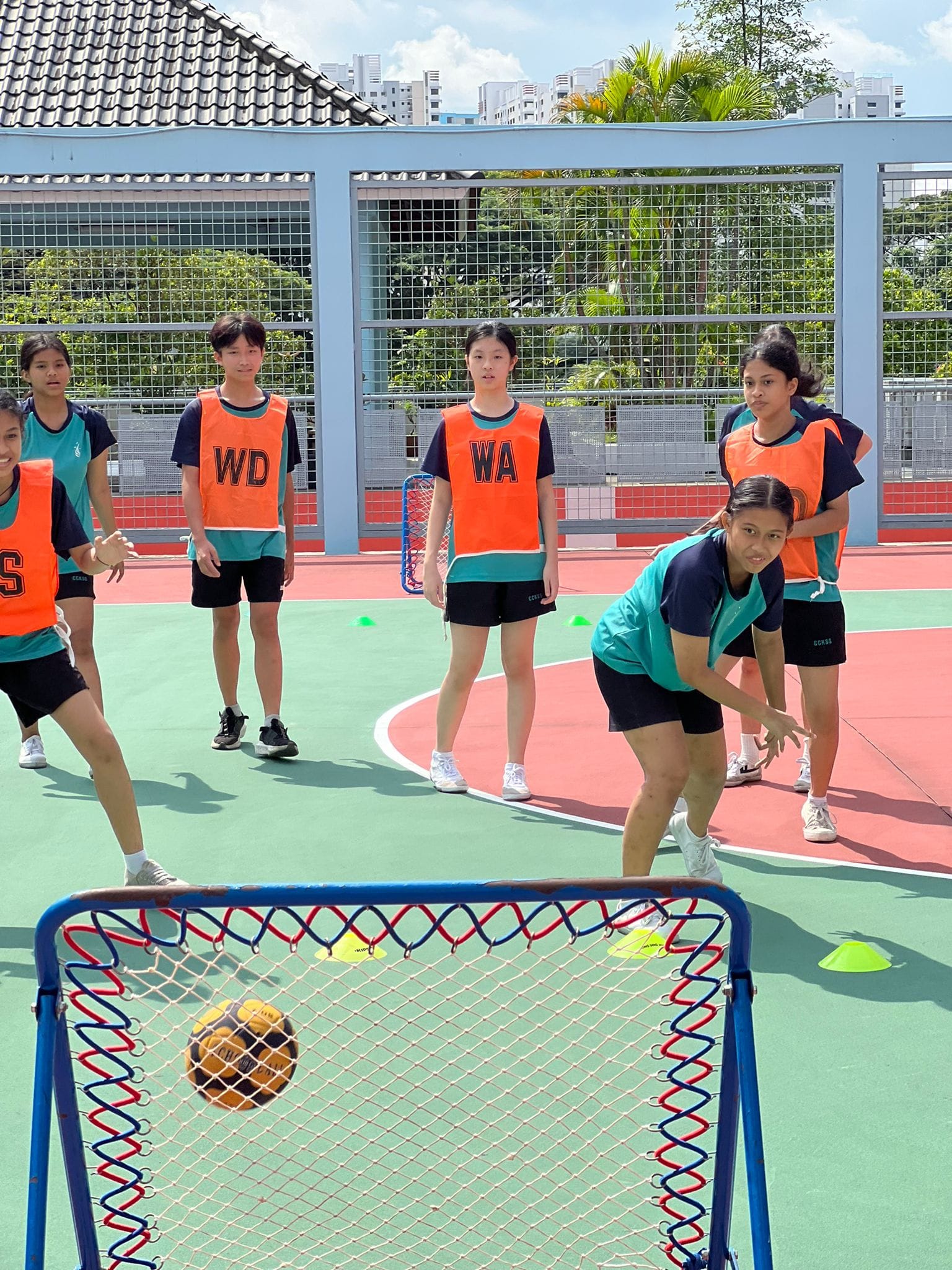 Students in orange bibs labeled WD and WA play netball on an outdoor court, with a ball near a small goal.