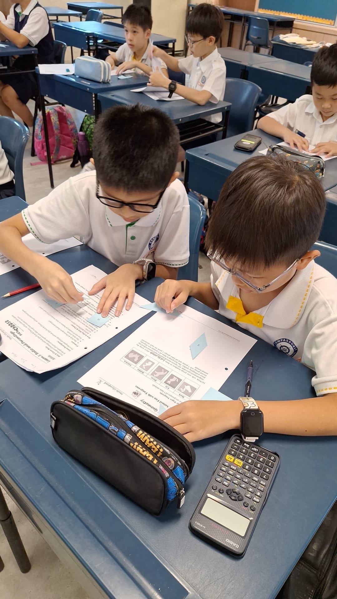 Two young students in white uniforms work together on a geometry worksheet at a blue desk with a Casio calculator nearby.