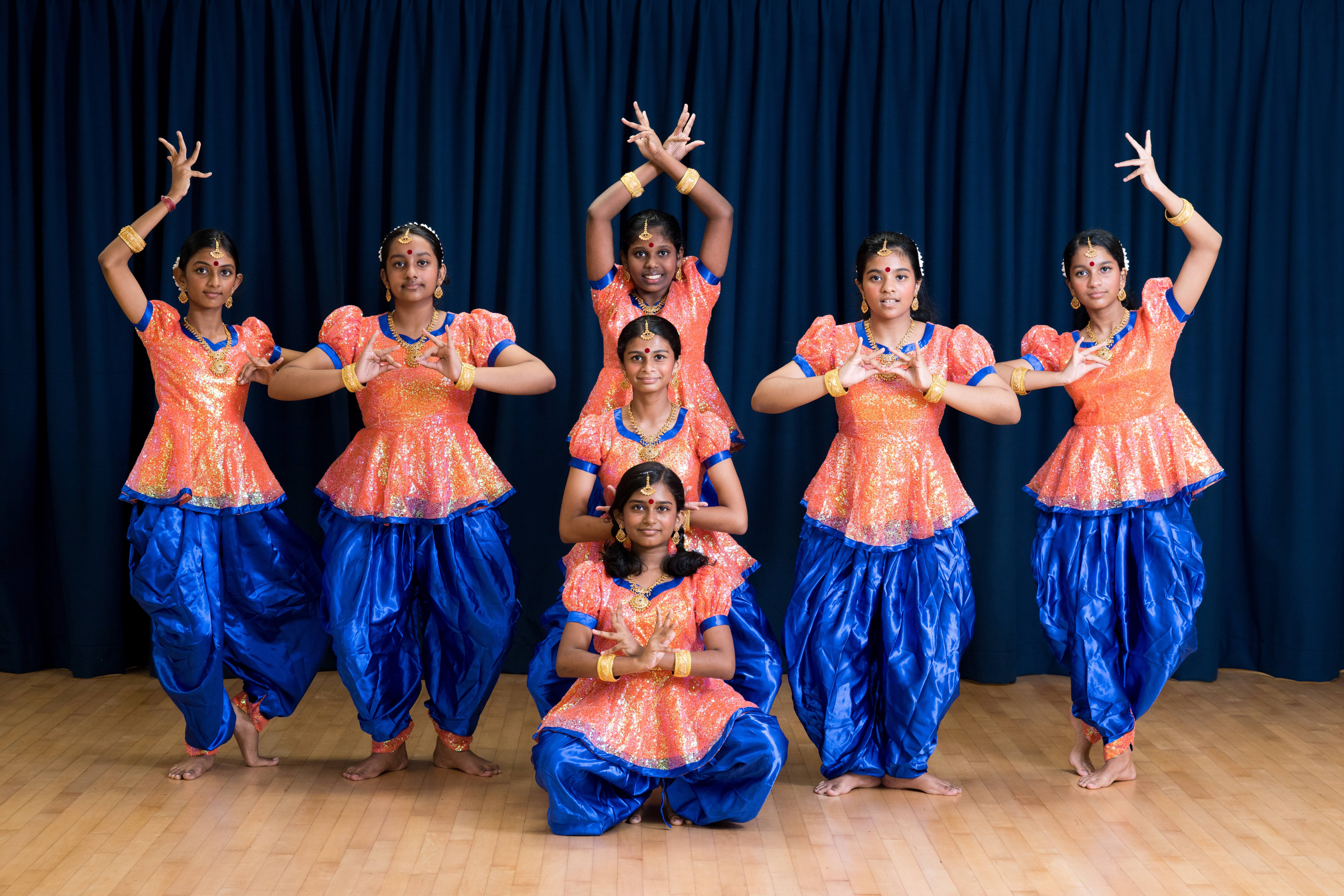 Six dancers in orange tops and blue pants pose with raised arms in front of a dark curtain.