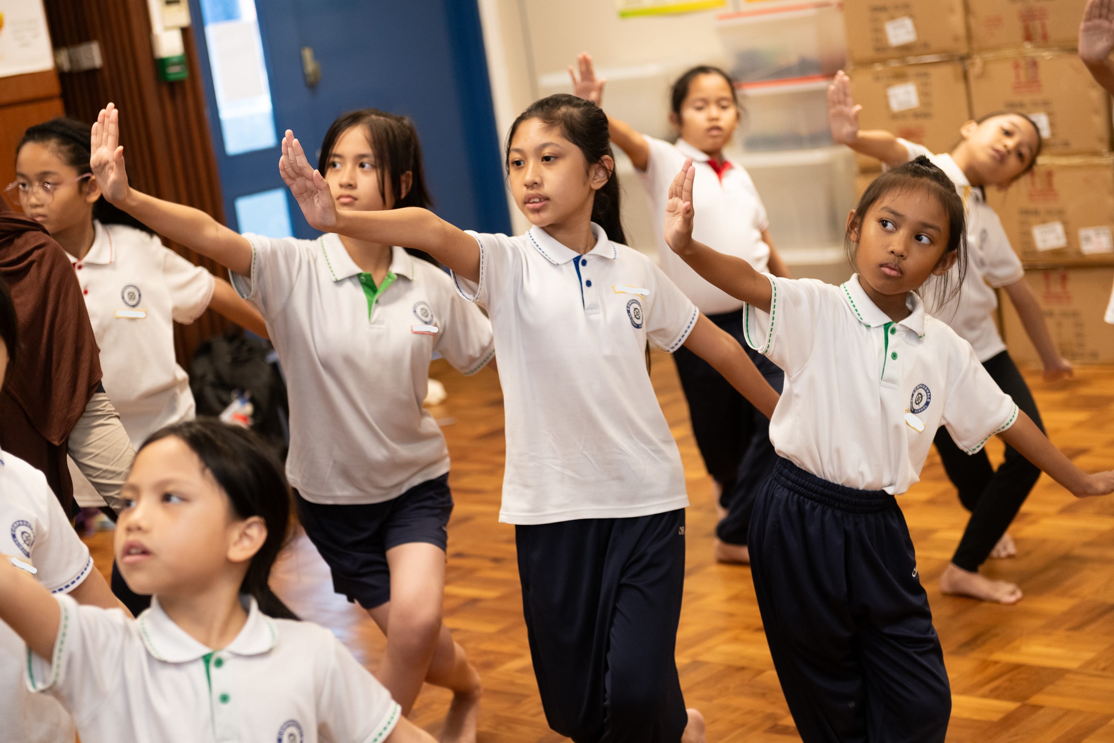 Several schoolgirls in white uniforms practice a dance routine with arms extended in an indoor wooden-floor room.