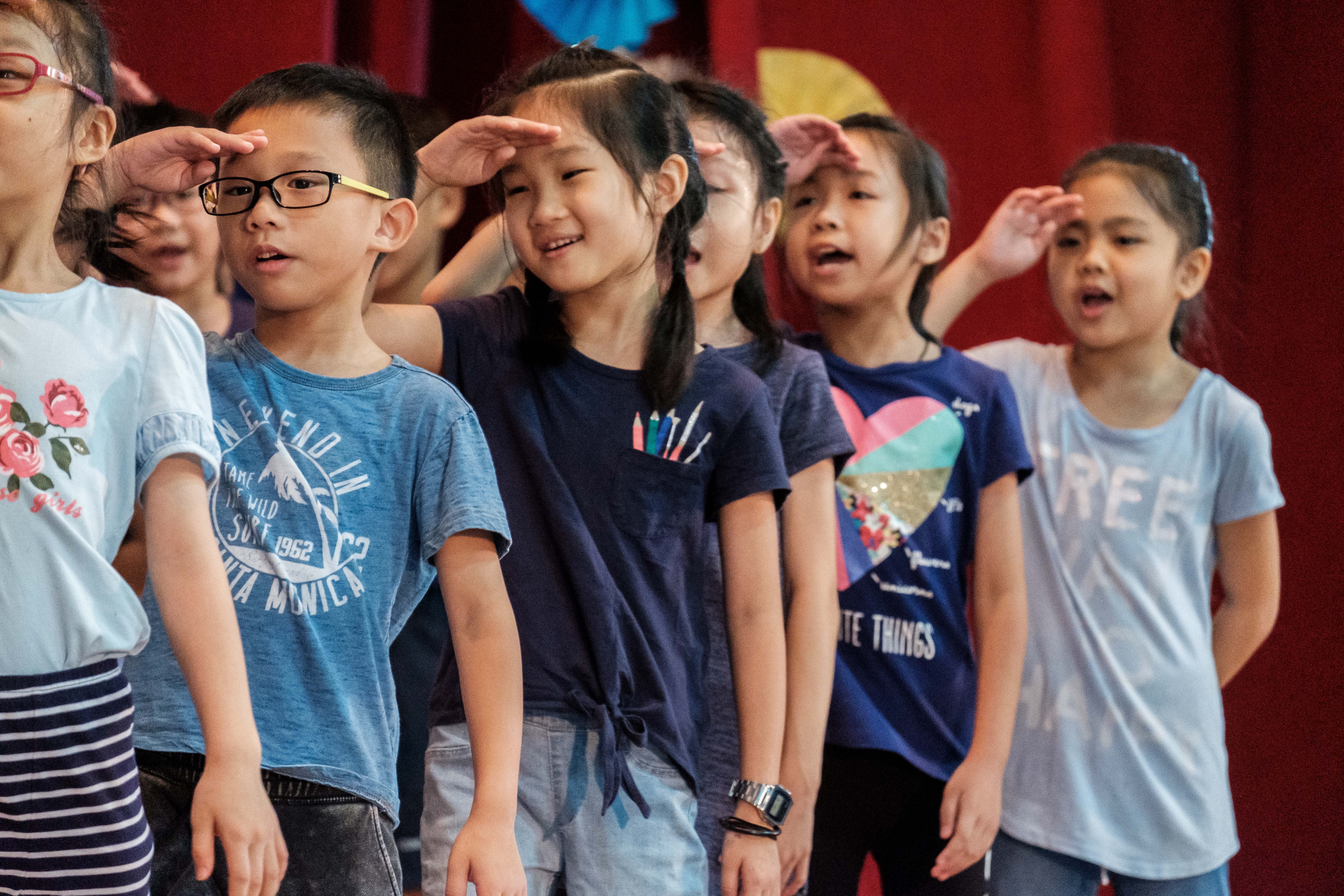 Several young children stand on a stage saluting with one hand raised to their foreheads before a red curtain.