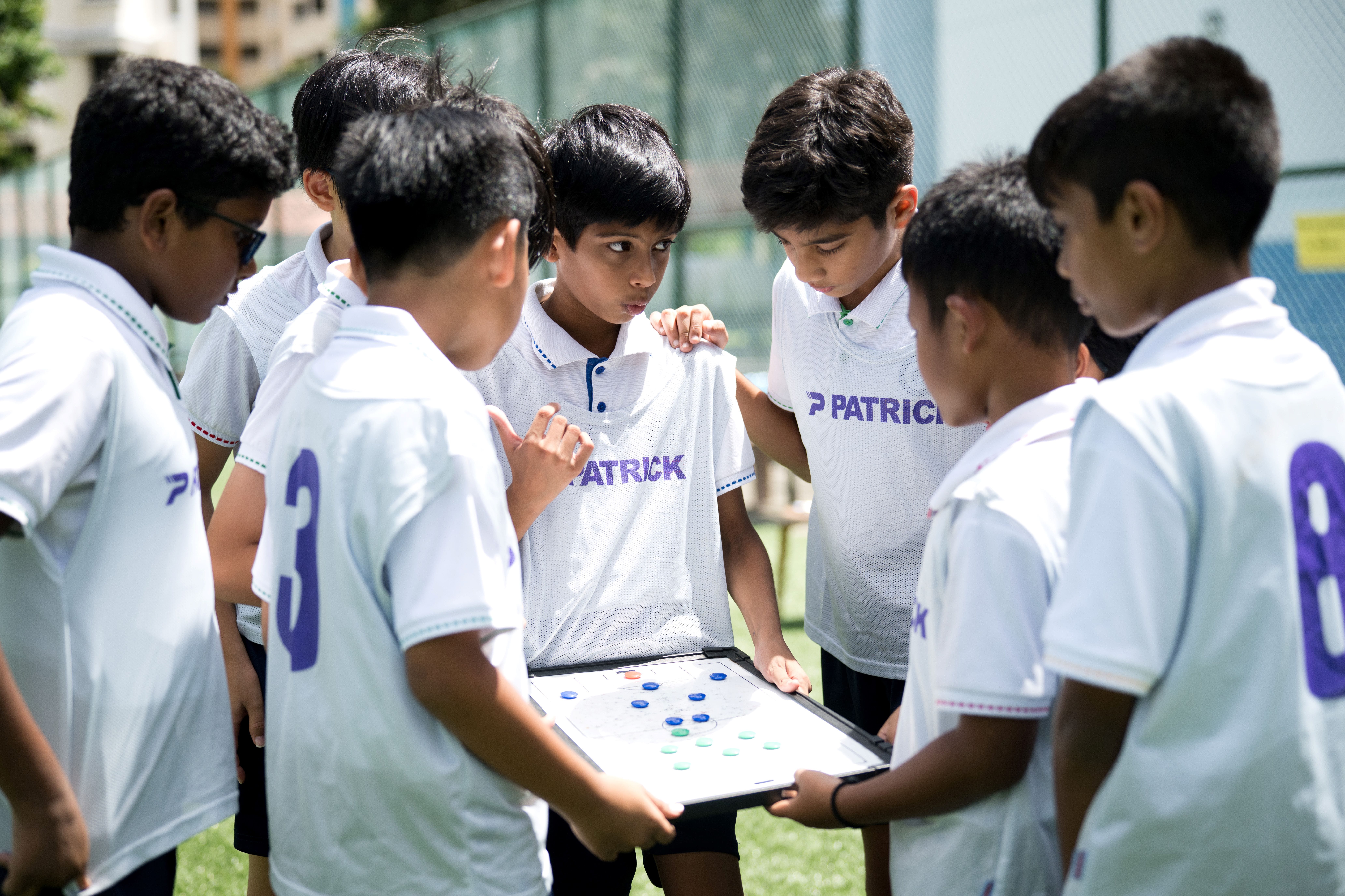 Boys in white soccer jerseys huddle around a coach holding a tactics board with blue and green markers.