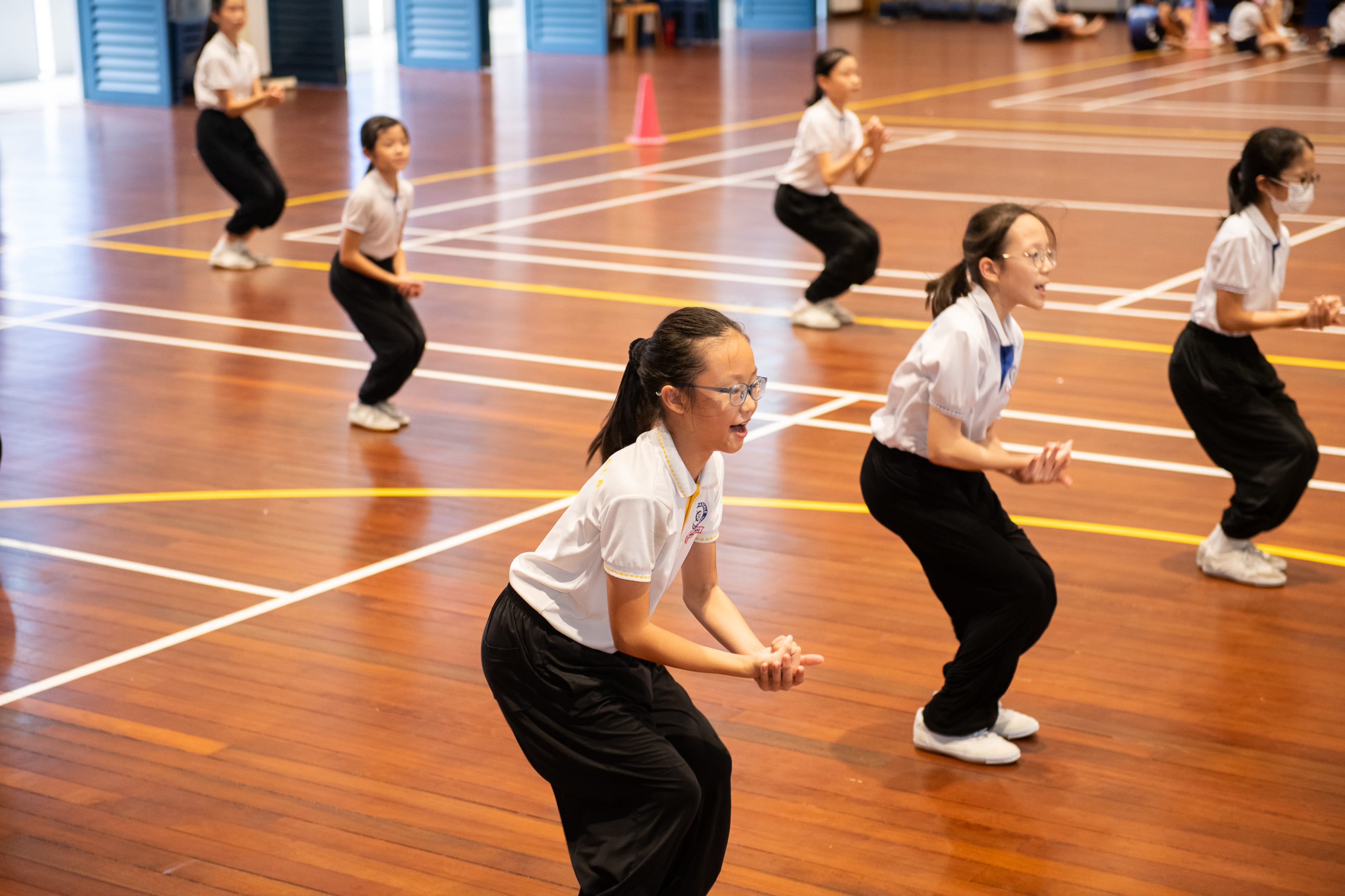 Students in white shirts and black pants practice squats on a gym floor with court lines.