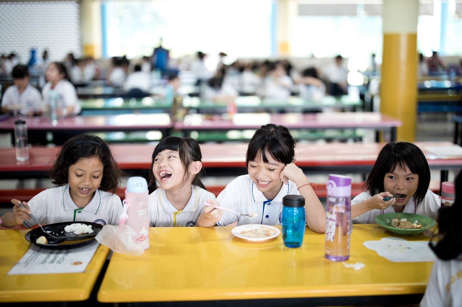 Four young girls in white school uniforms laugh and eat lunch together at a yellow cafeteria table.