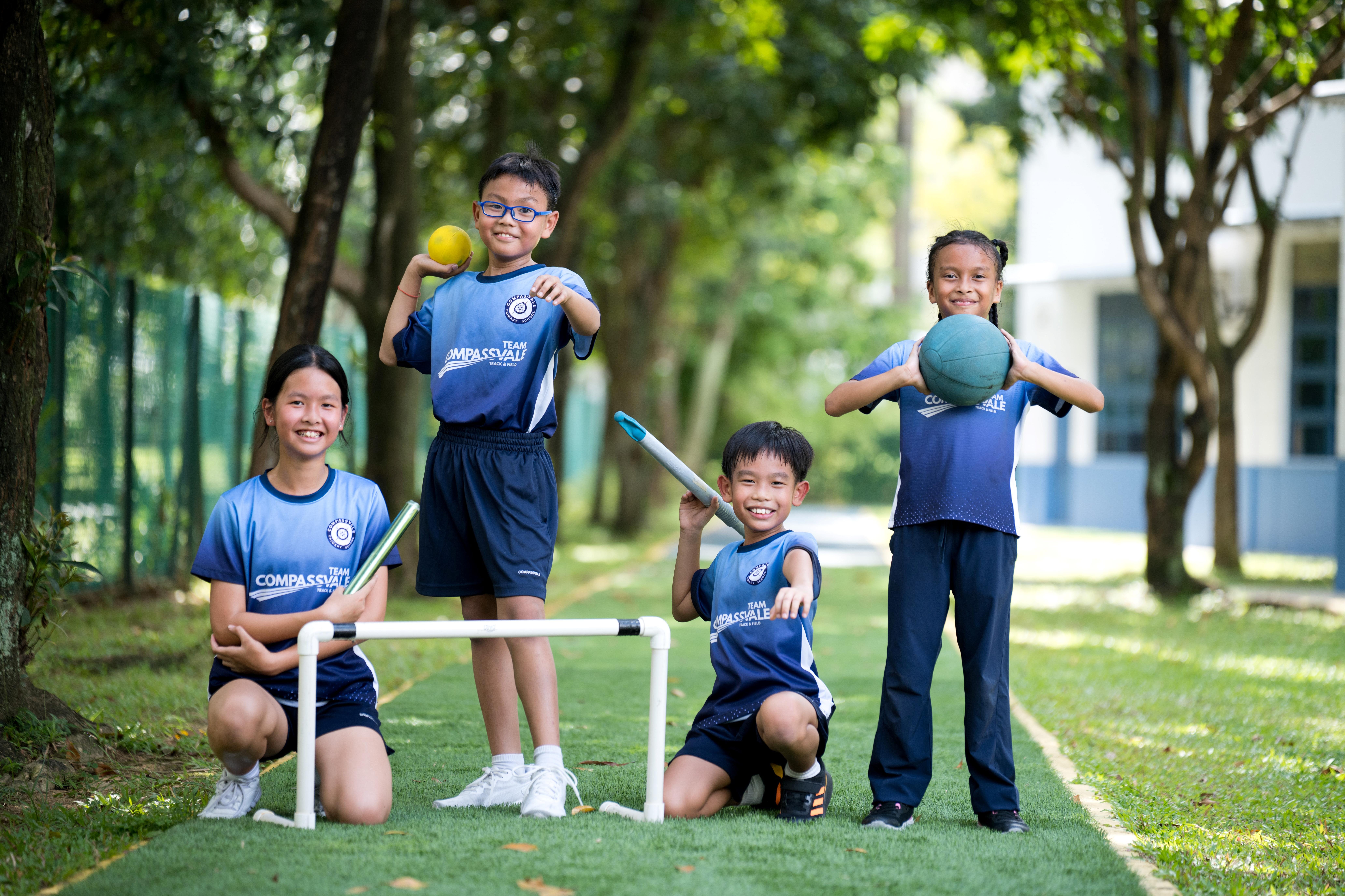 Four children in blue sports uniforms pose on a grassy path with balls, a bat, and a hurdle.