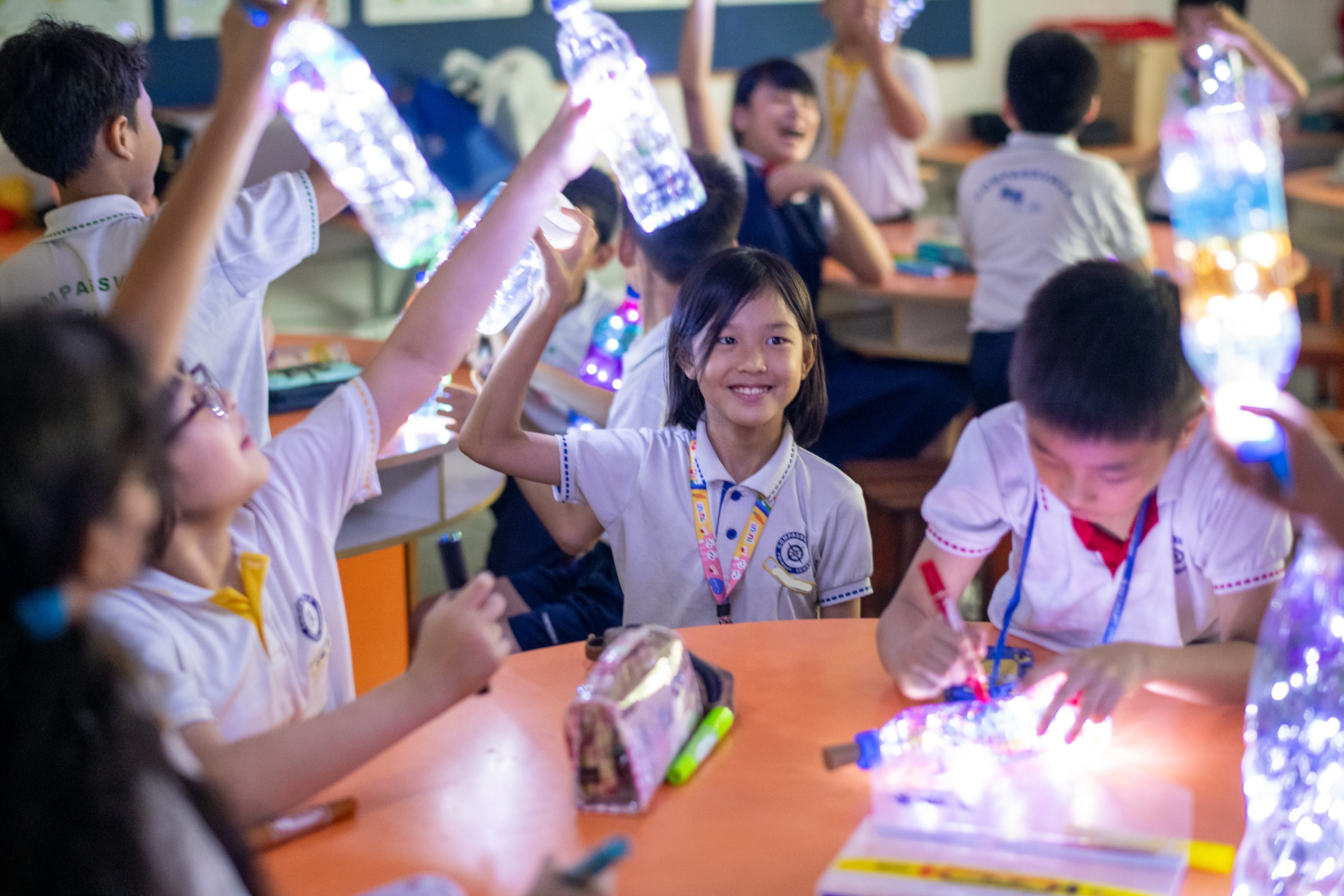 Children in school uniforms hold glowing light-filled plastic bottles during a classroom science activity.
