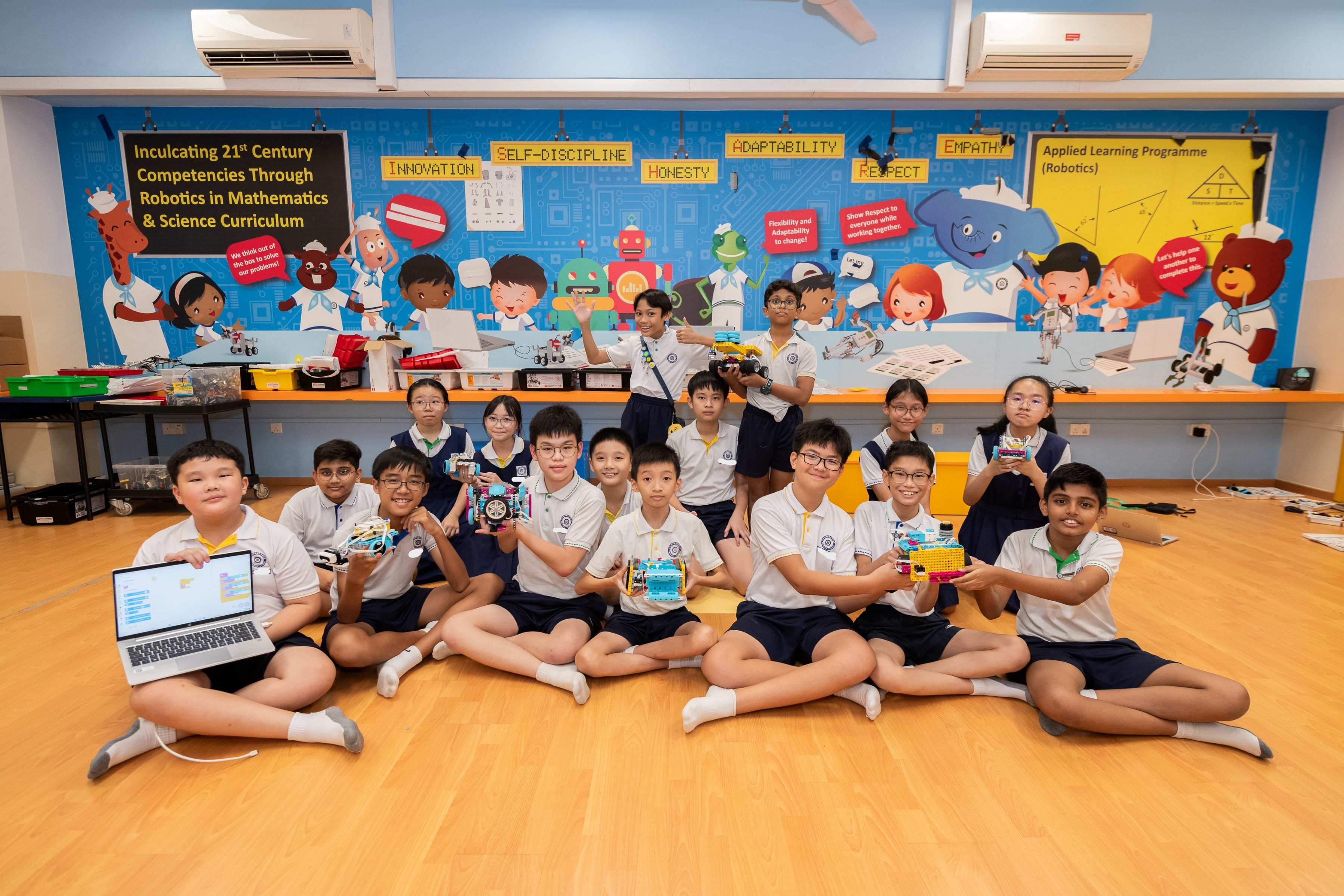 Students in school uniforms sit on a floor holding robots and a laptop in front of a colorful robotics classroom mural.
