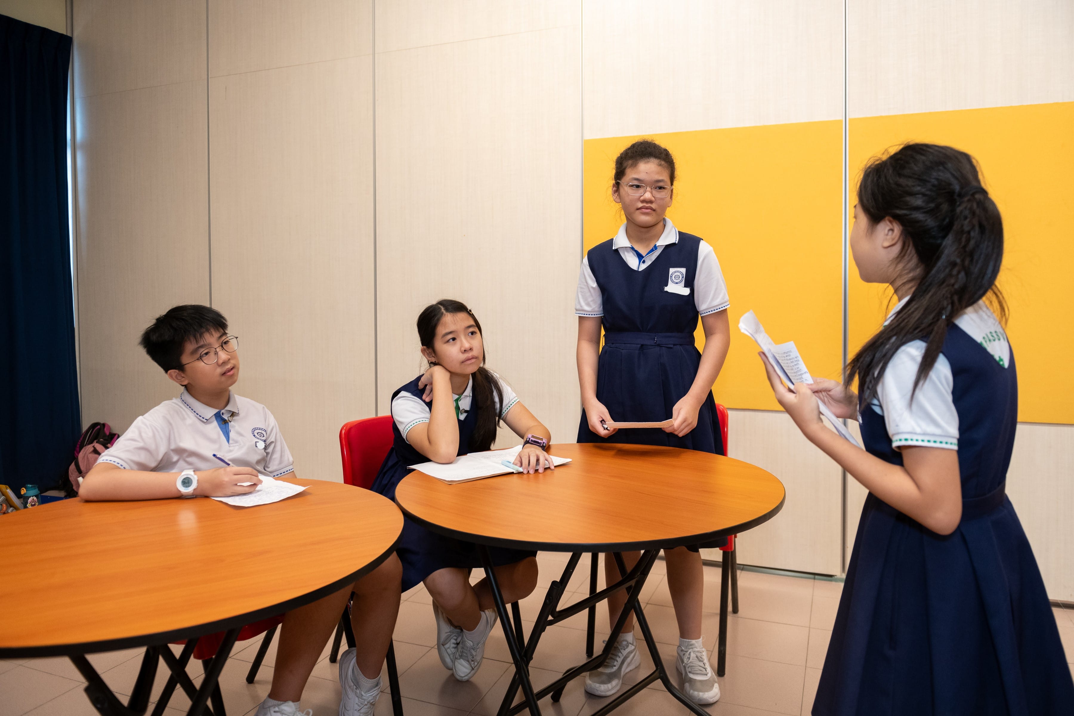 Four students in navy and white uniforms gather around round tables, two standing and two seated, holding papers.