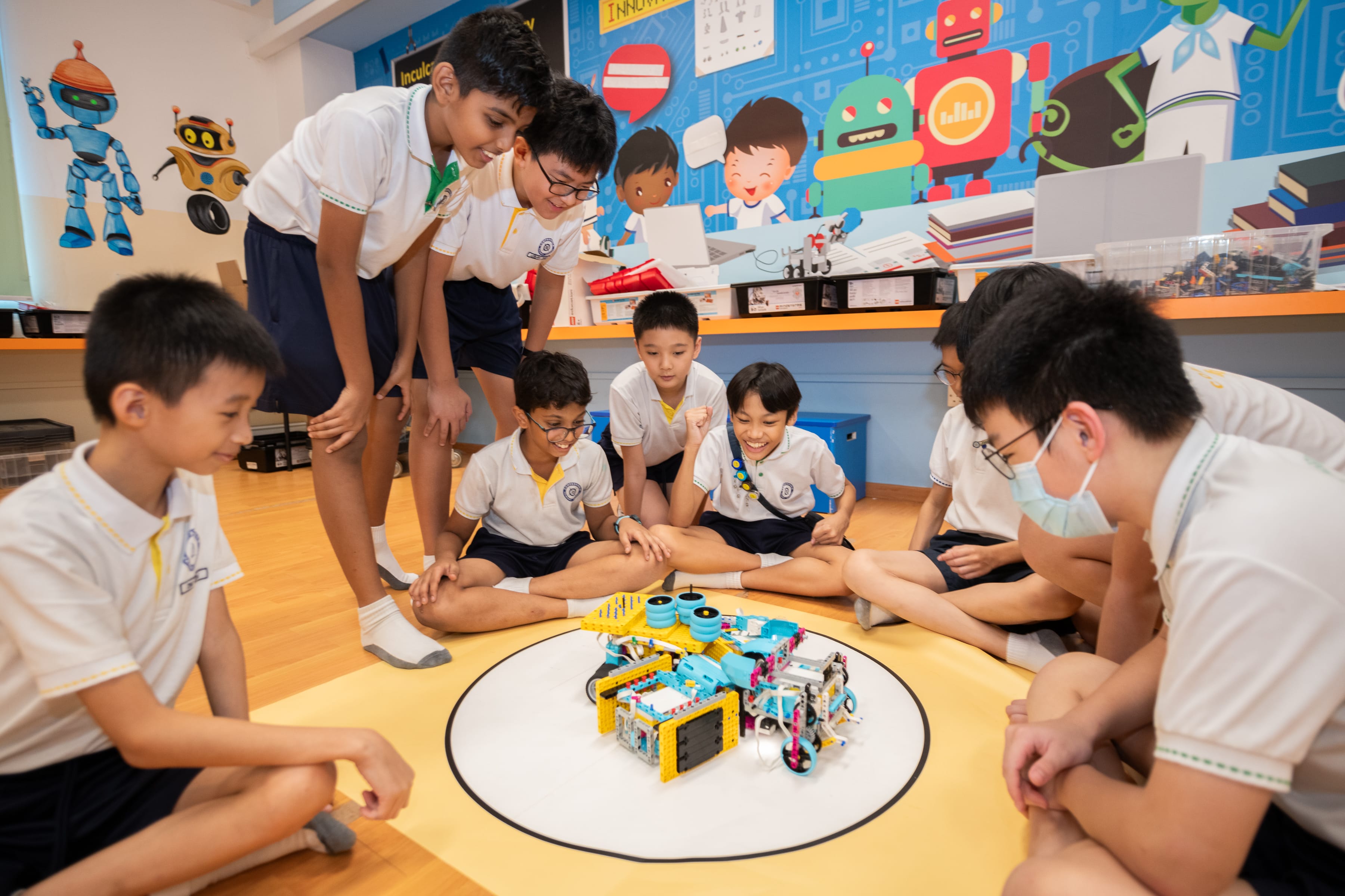 Group of students in uniform sit in a circle watching a small robot on a floor mat in a classroom.