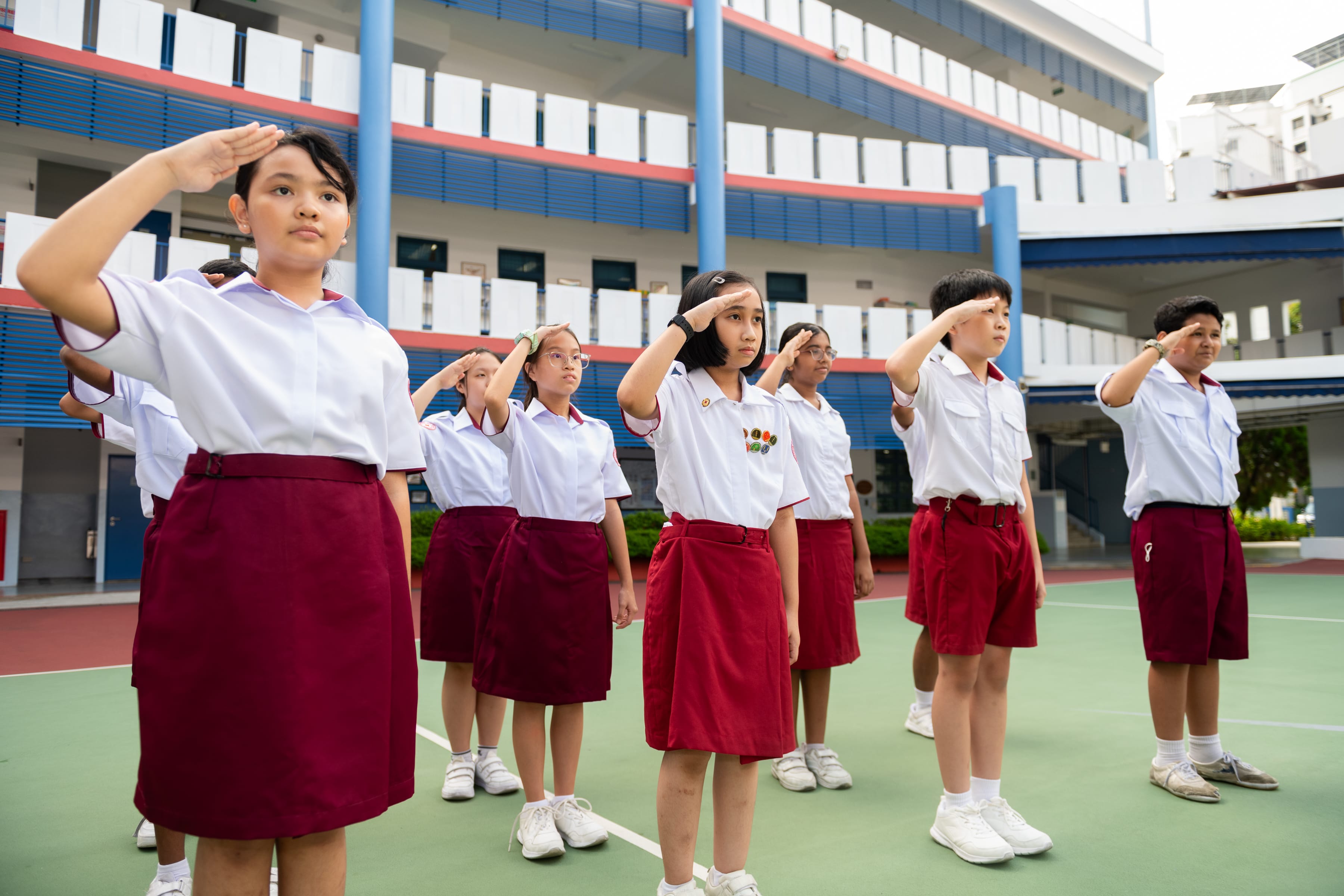 Students in white and red uniforms saluting in rows on a school courtyard with a multi-storey building behind them.
