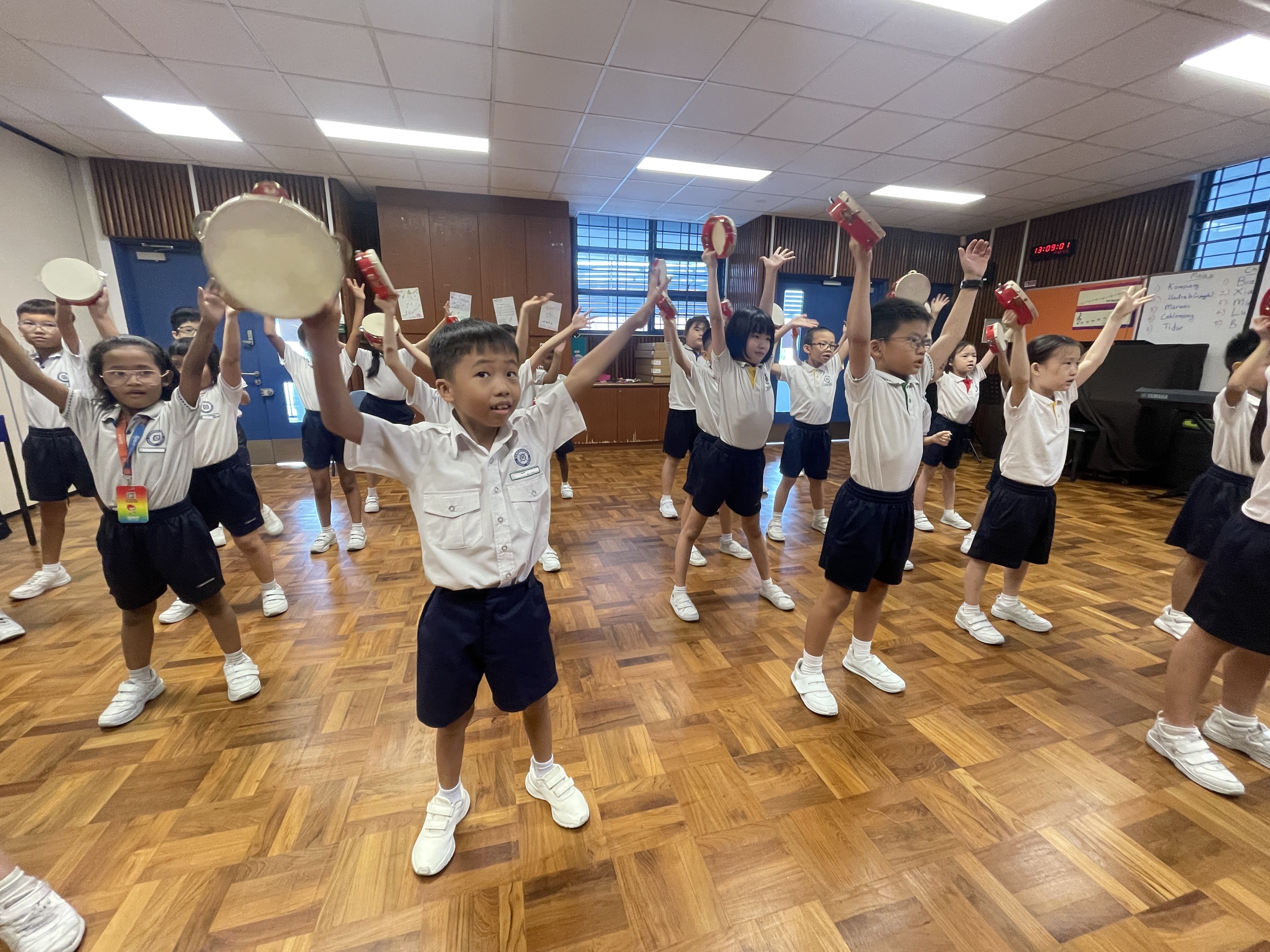 Young students in school uniforms raise tambourines and percussion instruments overhead during a music class indoors.