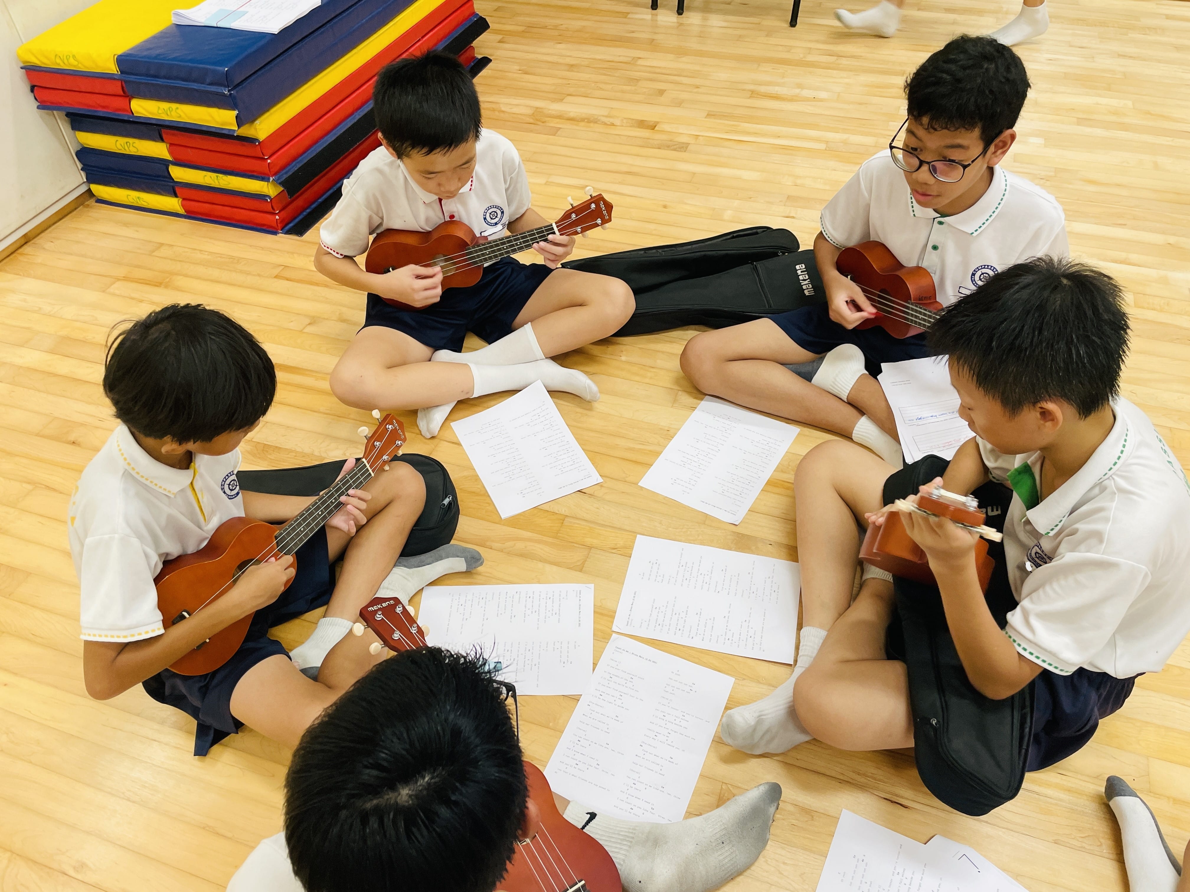 Four school children in uniforms sit on a wooden floor playing ukuleles with sheet music papers spread around them.