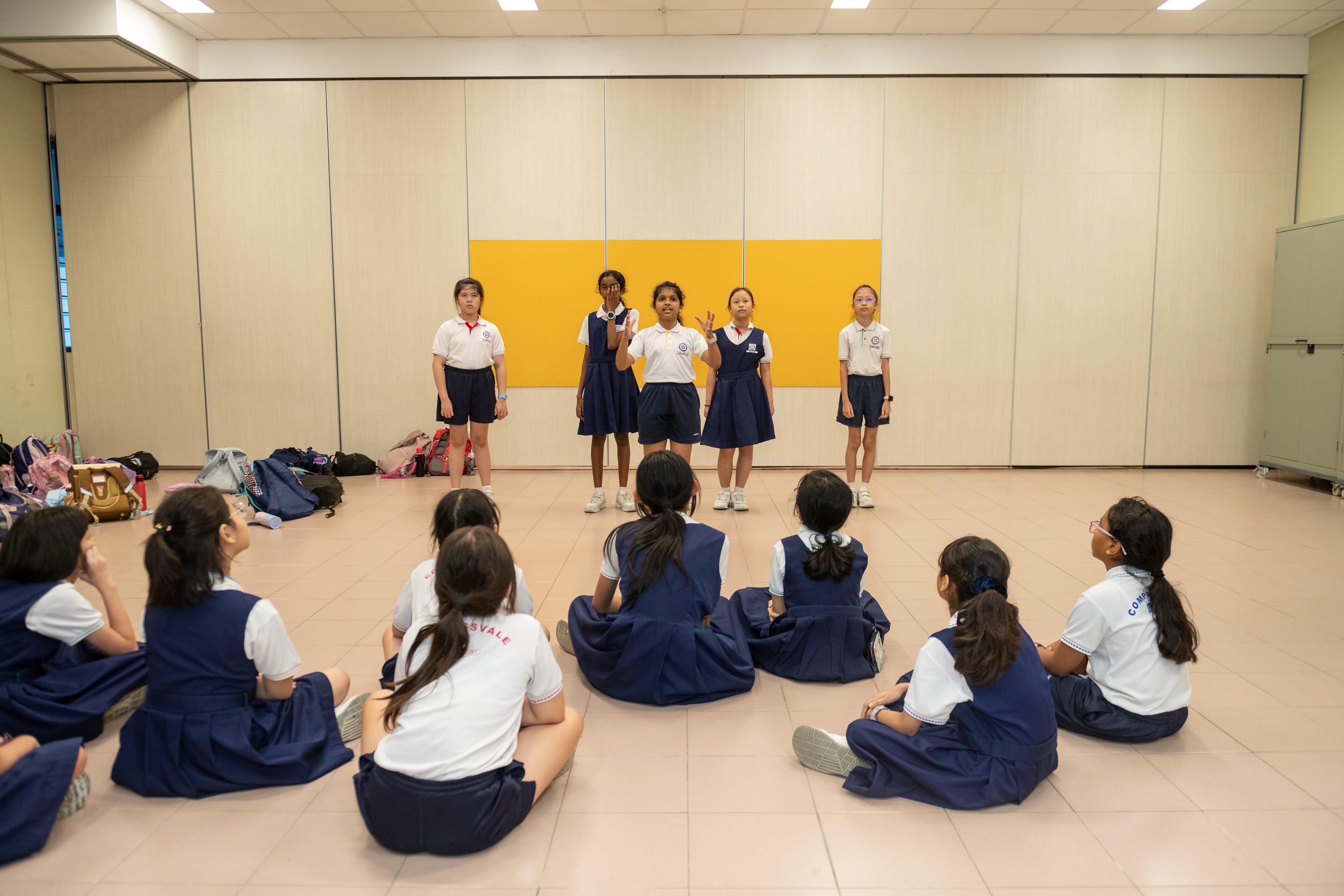 Five students stand presenting to classmates seated on the floor in a school hall with bags along the wall.