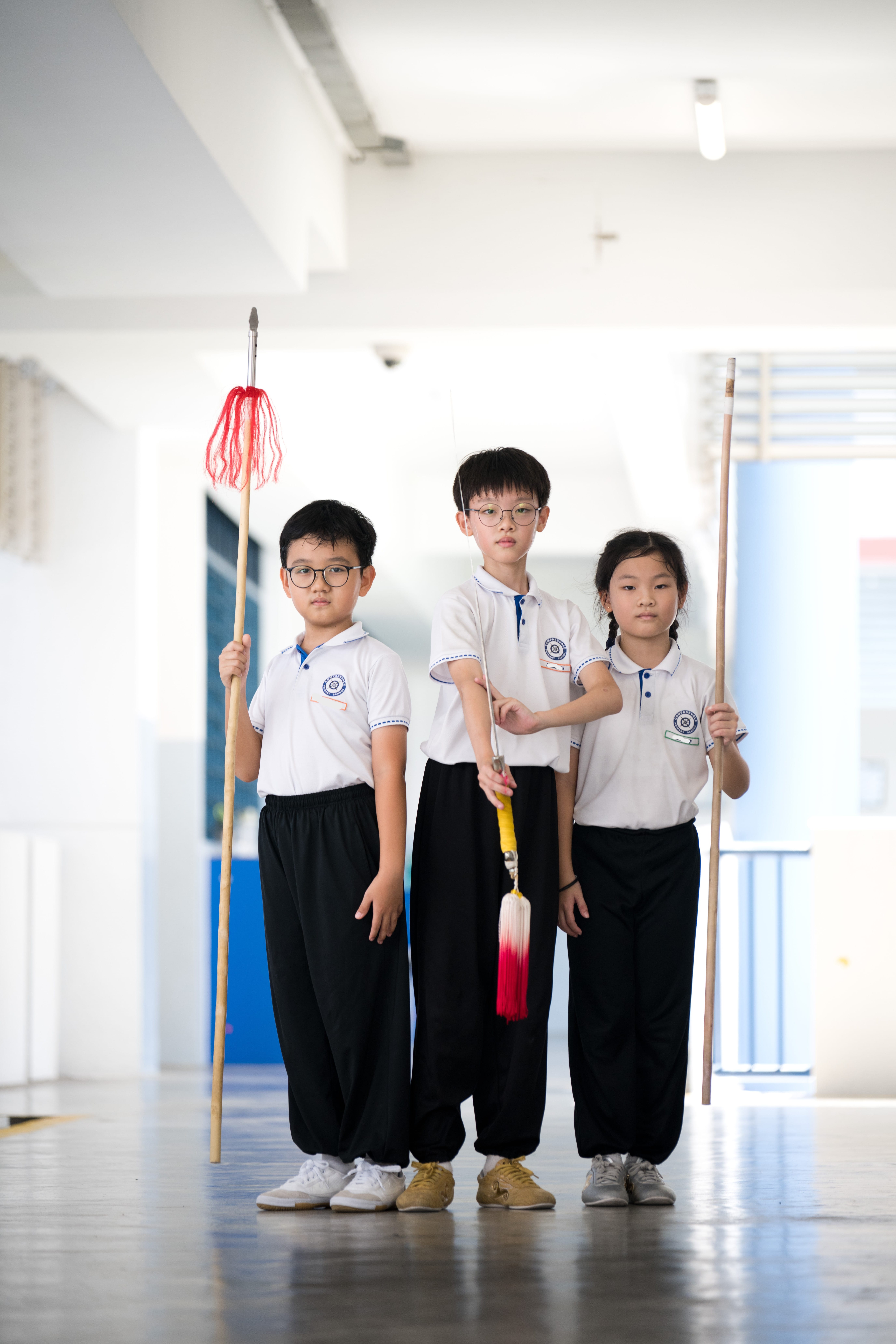 Three schoolchildren in uniforms stand in a corridor holding long sticks with red tassels.