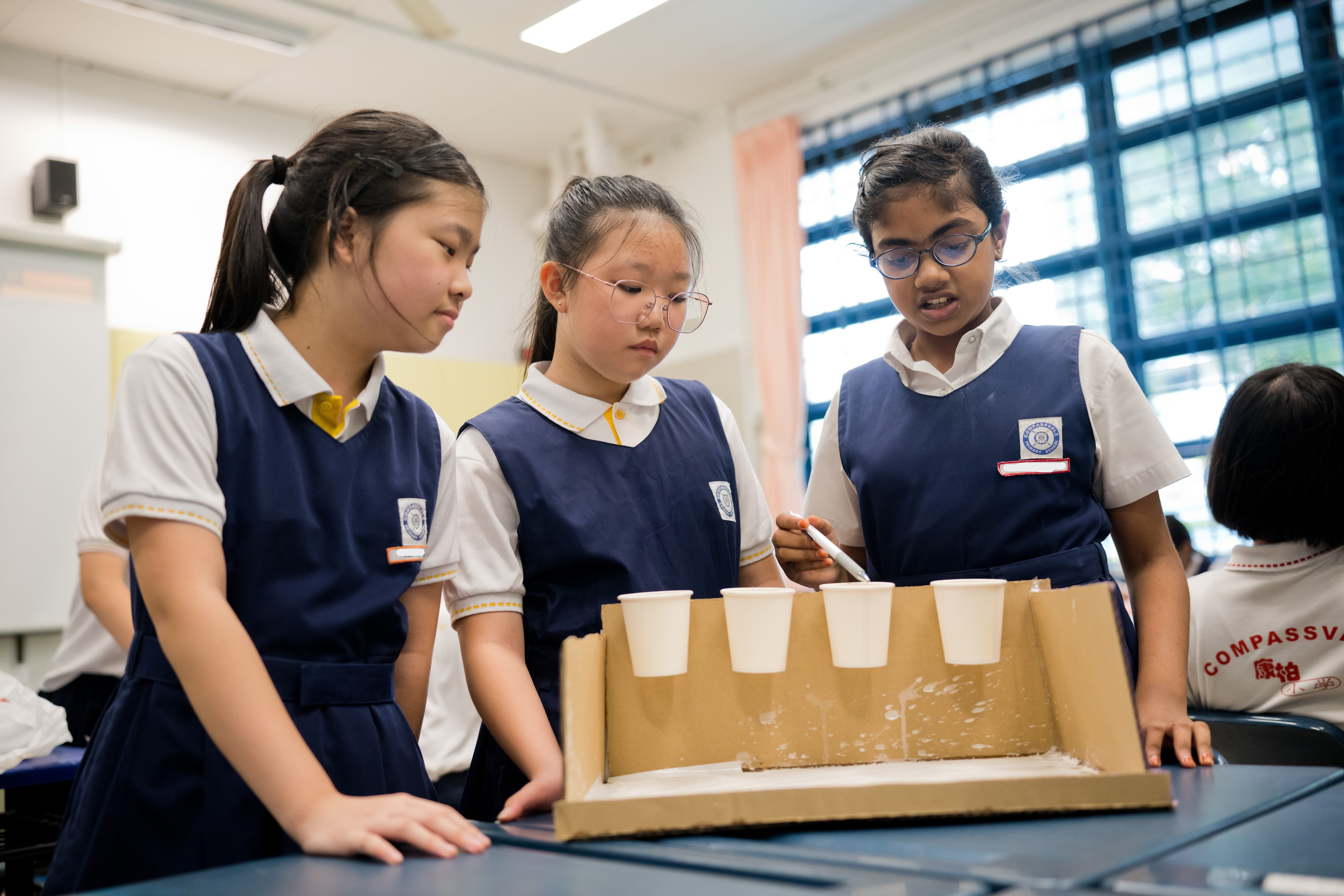 Three schoolgirls in uniforms look at paper cups lined on a cardboard tray during a classroom activity.