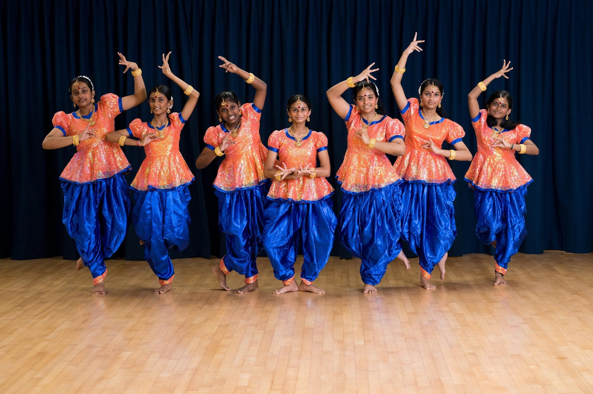 Seven girls in orange tops and blue harem pants perform a traditional Indian dance on a wooden stage.