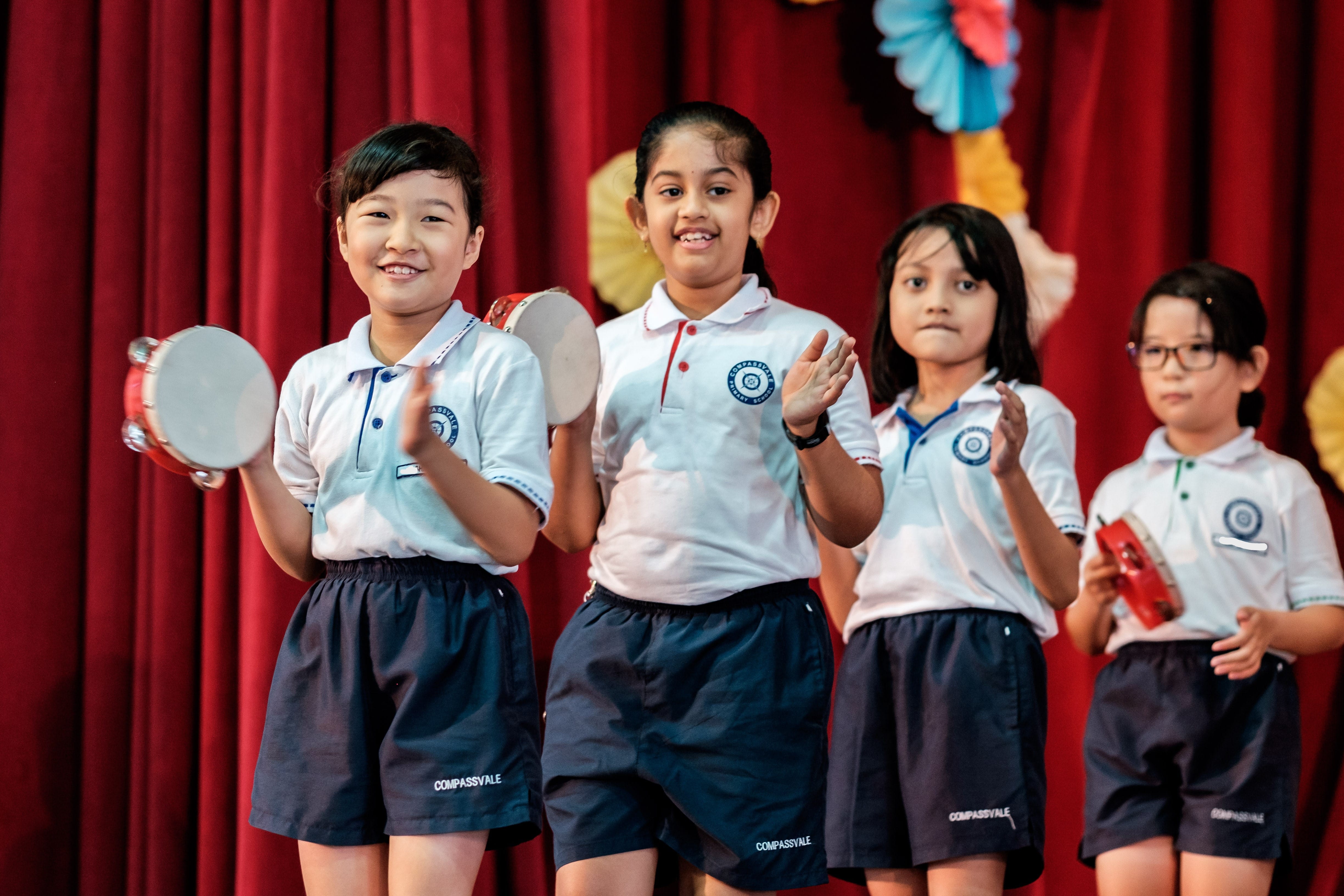 Four young students in Compassvale school uniforms perform on stage, smiling and holding tambourines before a red curtain.