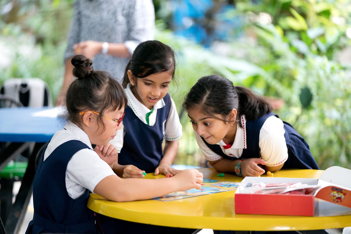 Three young girls in school uniforms play a board game with dice at a yellow outdoor table.