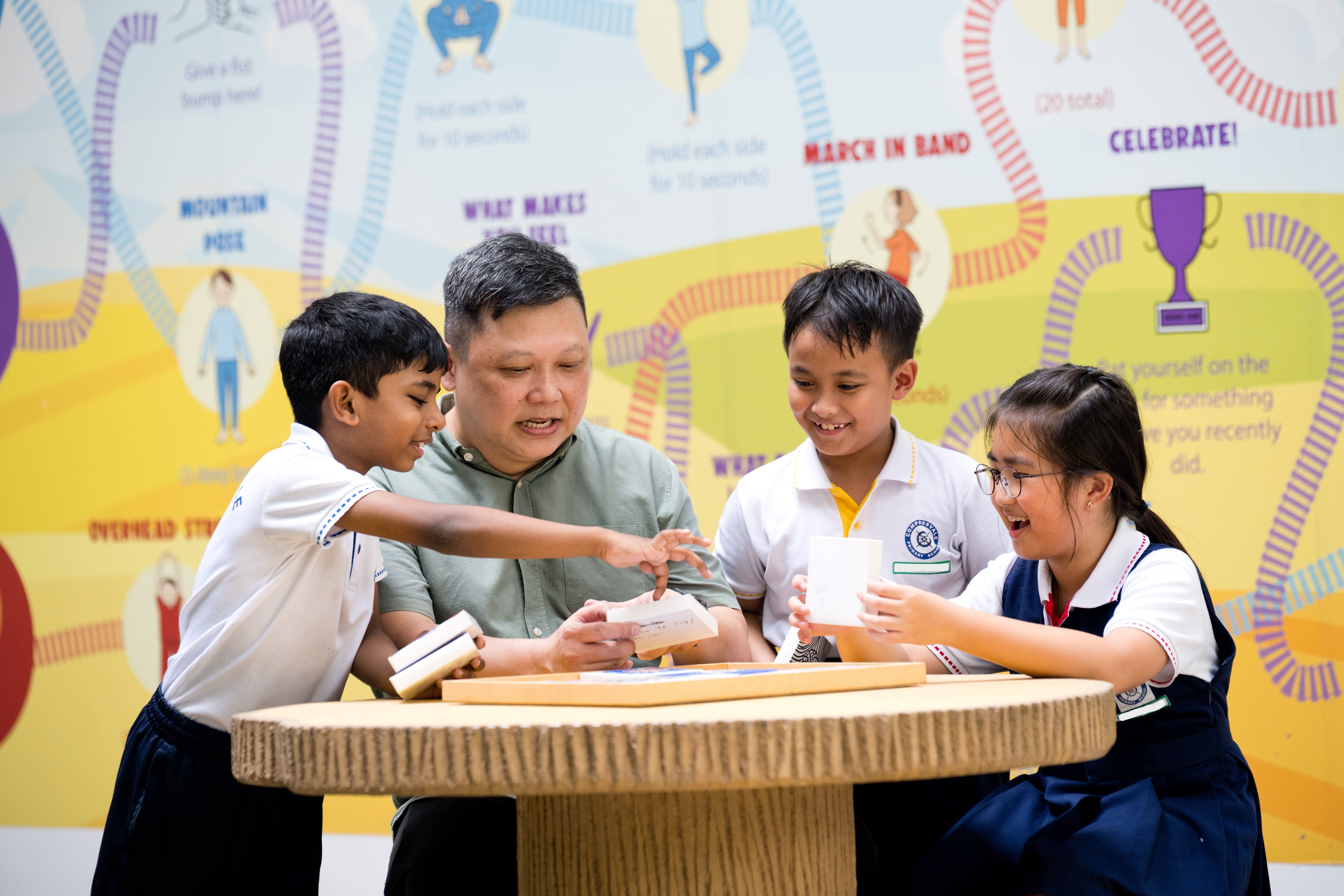 Teacher and three students at a round table, smiling and holding paper cards in a colorful classroom.